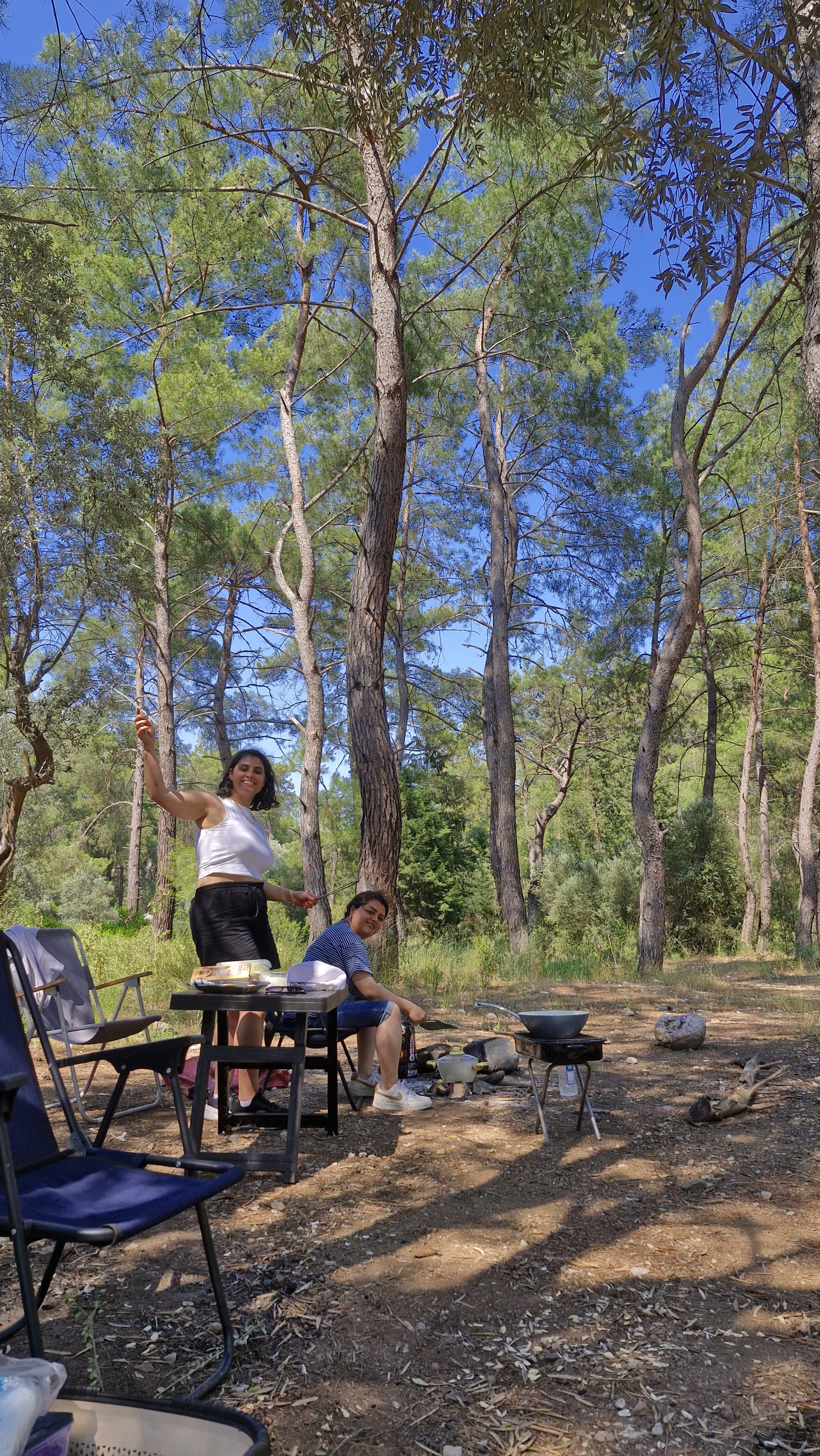 Two women stand and sit beside a table, folding chairs and cooking equipment among tall pine trees.