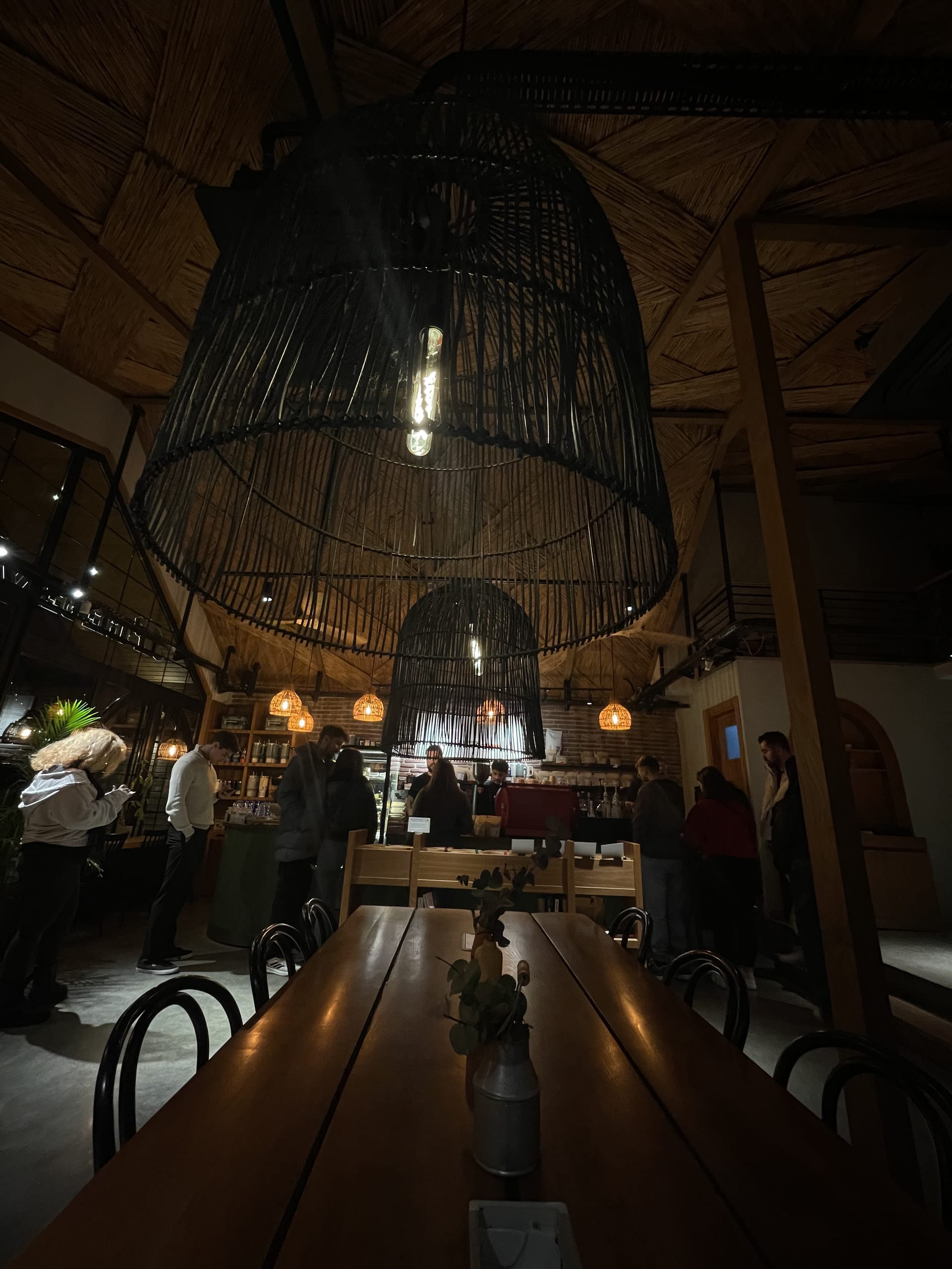 Large woven pendant lights hang above wooden tables in a warmly lit café filled with visitors.