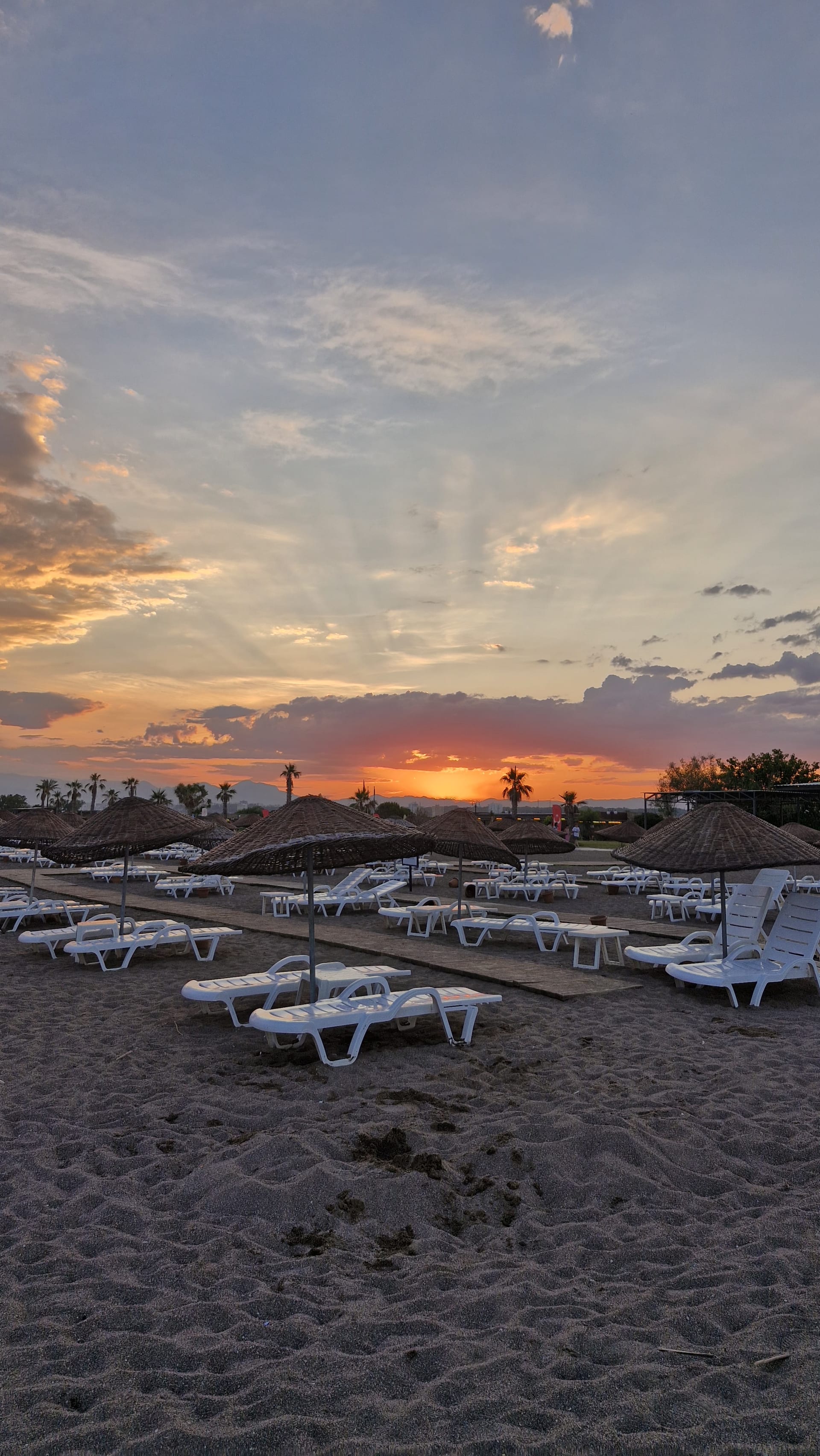 White sun loungers and straw parasols stand on the sand beneath an orange and pink sunset.