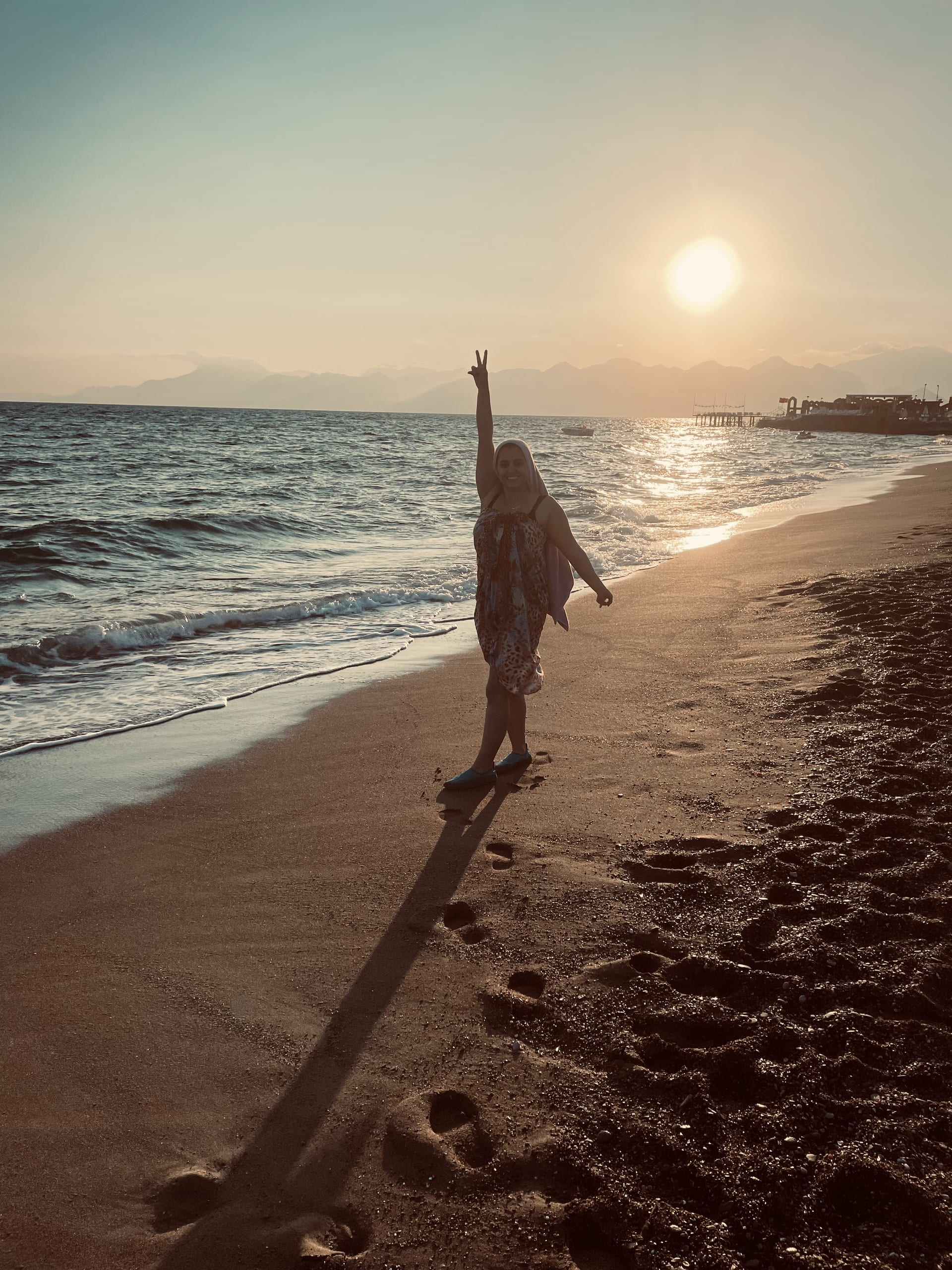 A woman stands at the water’s edge with one arm raised as the low sun reflects across the sea.