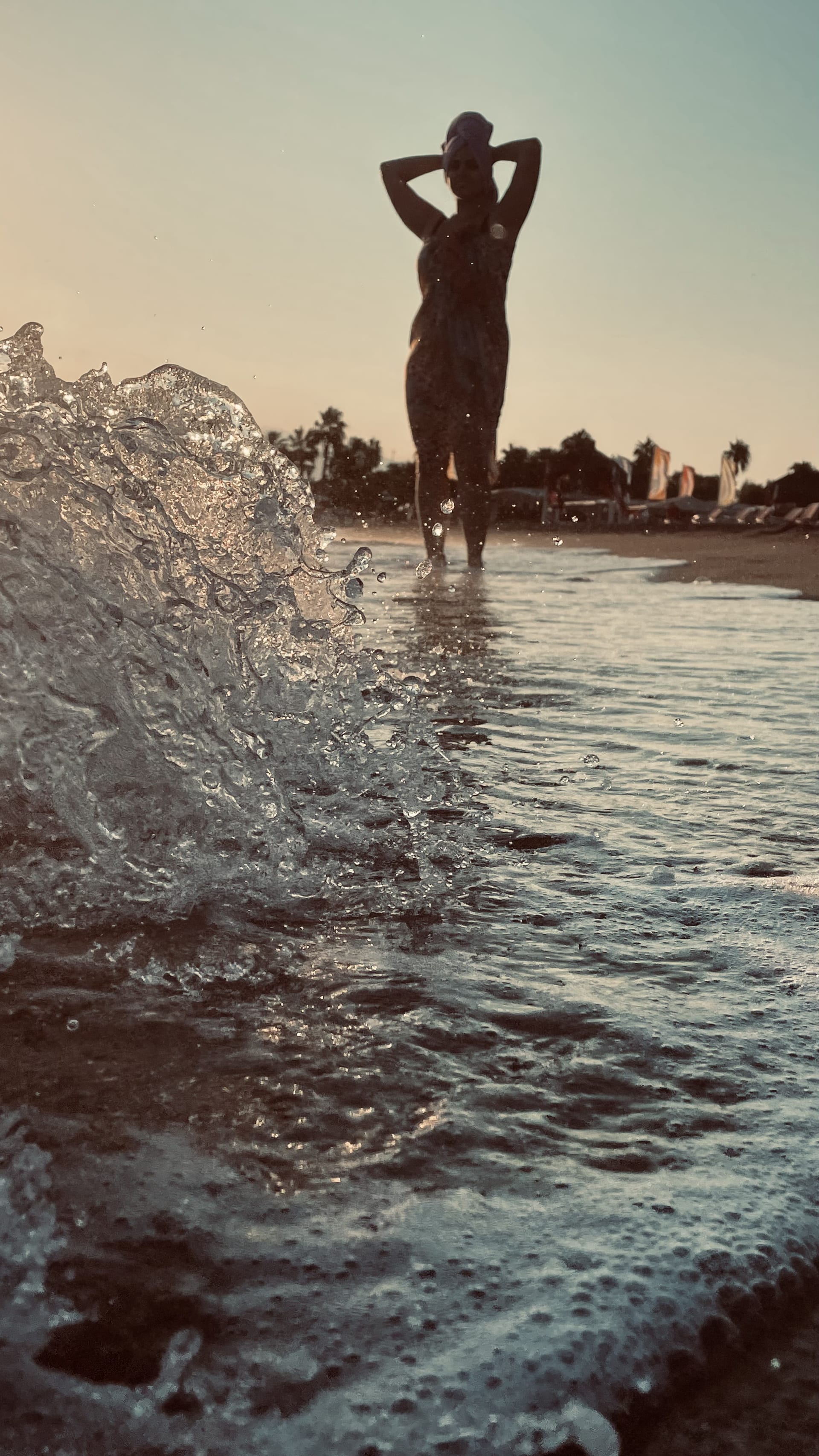 A small wave rises in the foreground while a woman stands silhouetted in the shallow water.