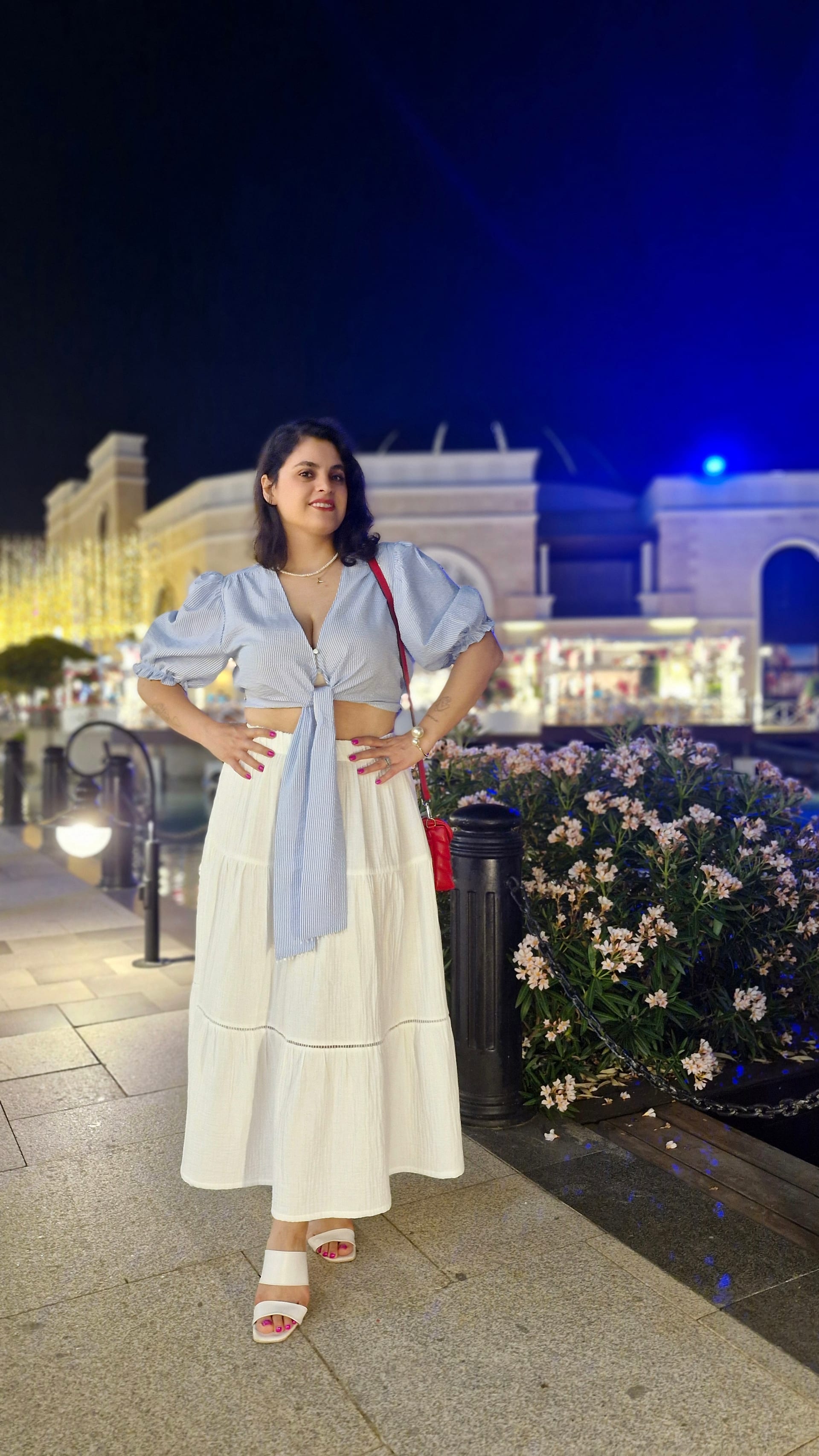 A woman poses beside flowers and decorative lamps with illuminated buildings behind her.