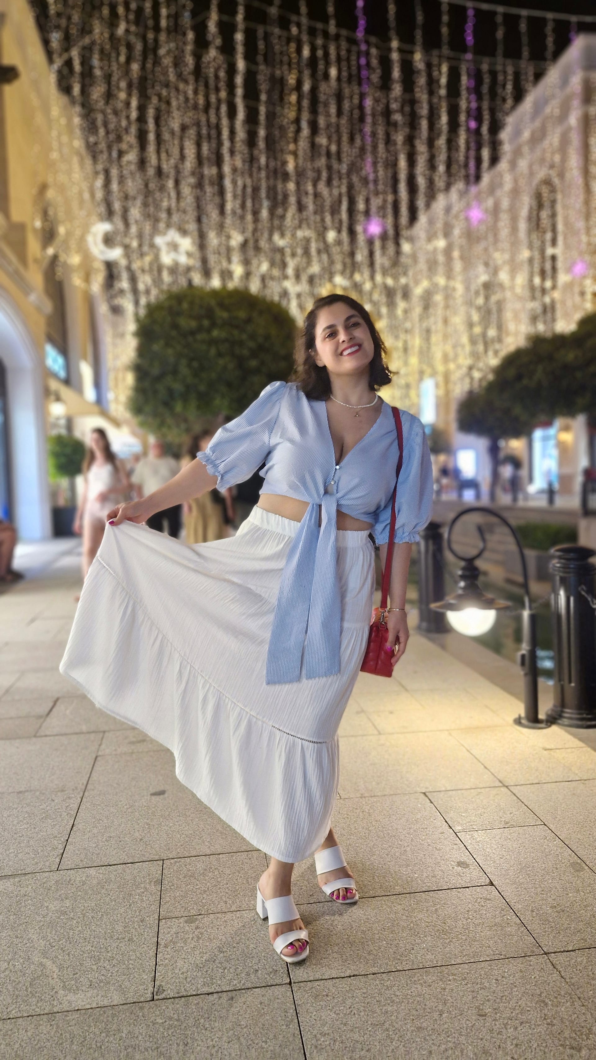 A woman poses beneath dense strands of white and purple lights along a pedestrian street.