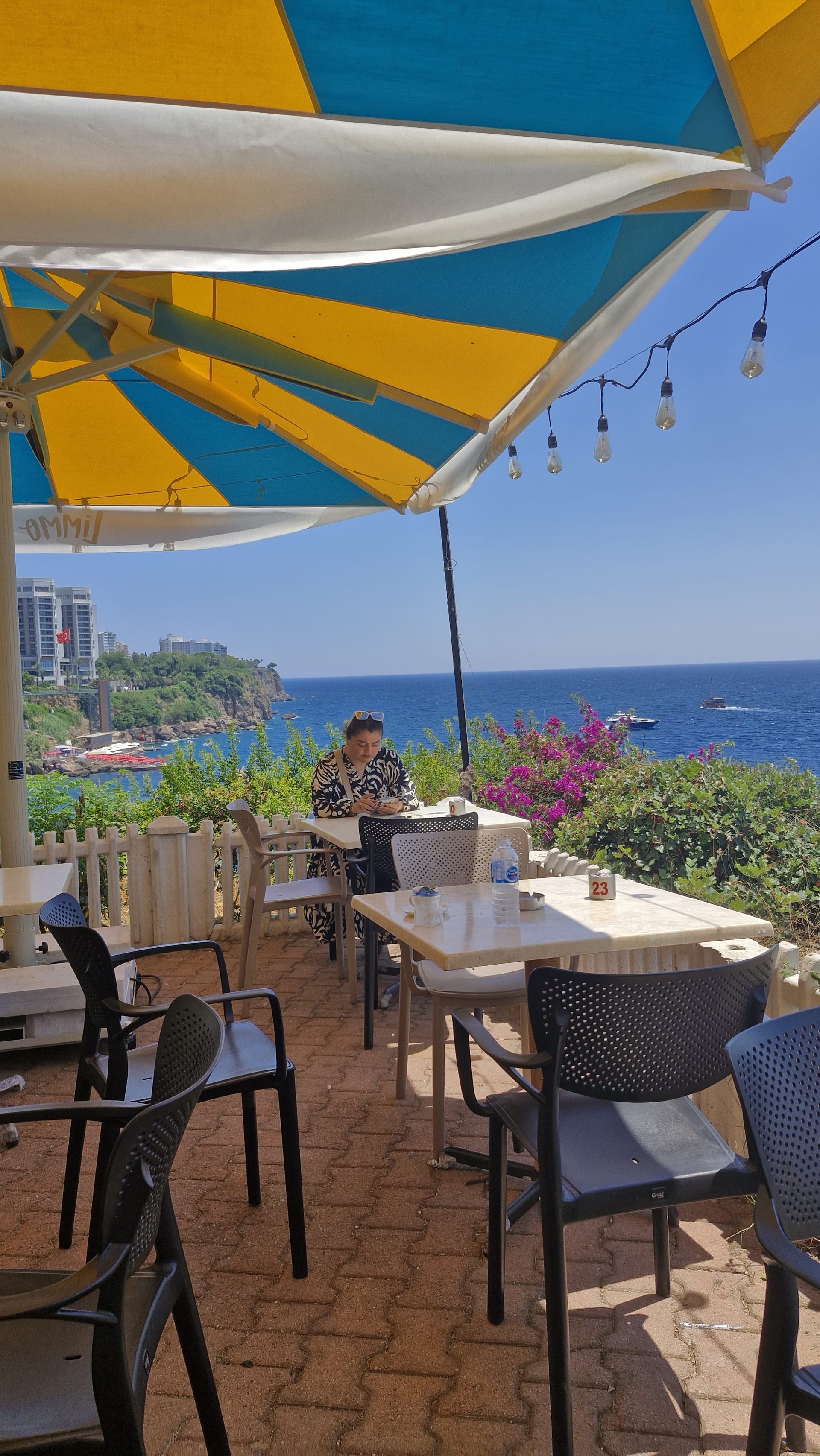 Café tables sit beneath yellow-and-blue awnings overlooking cliffs, blue sea and flowering plants.