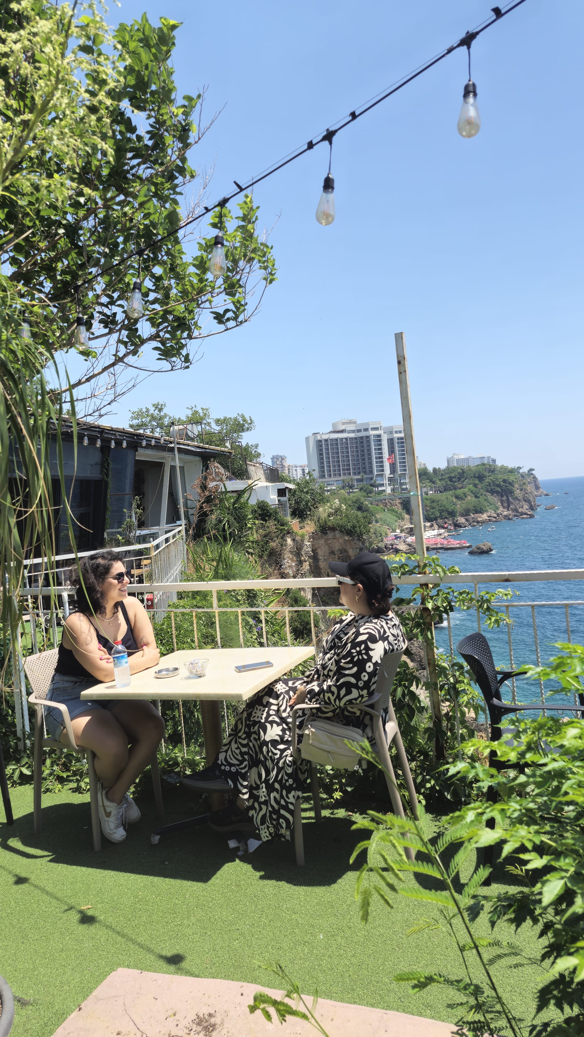 Two women sit together at a cliffside café table with greenery, blue water and rocky coastline behind them.