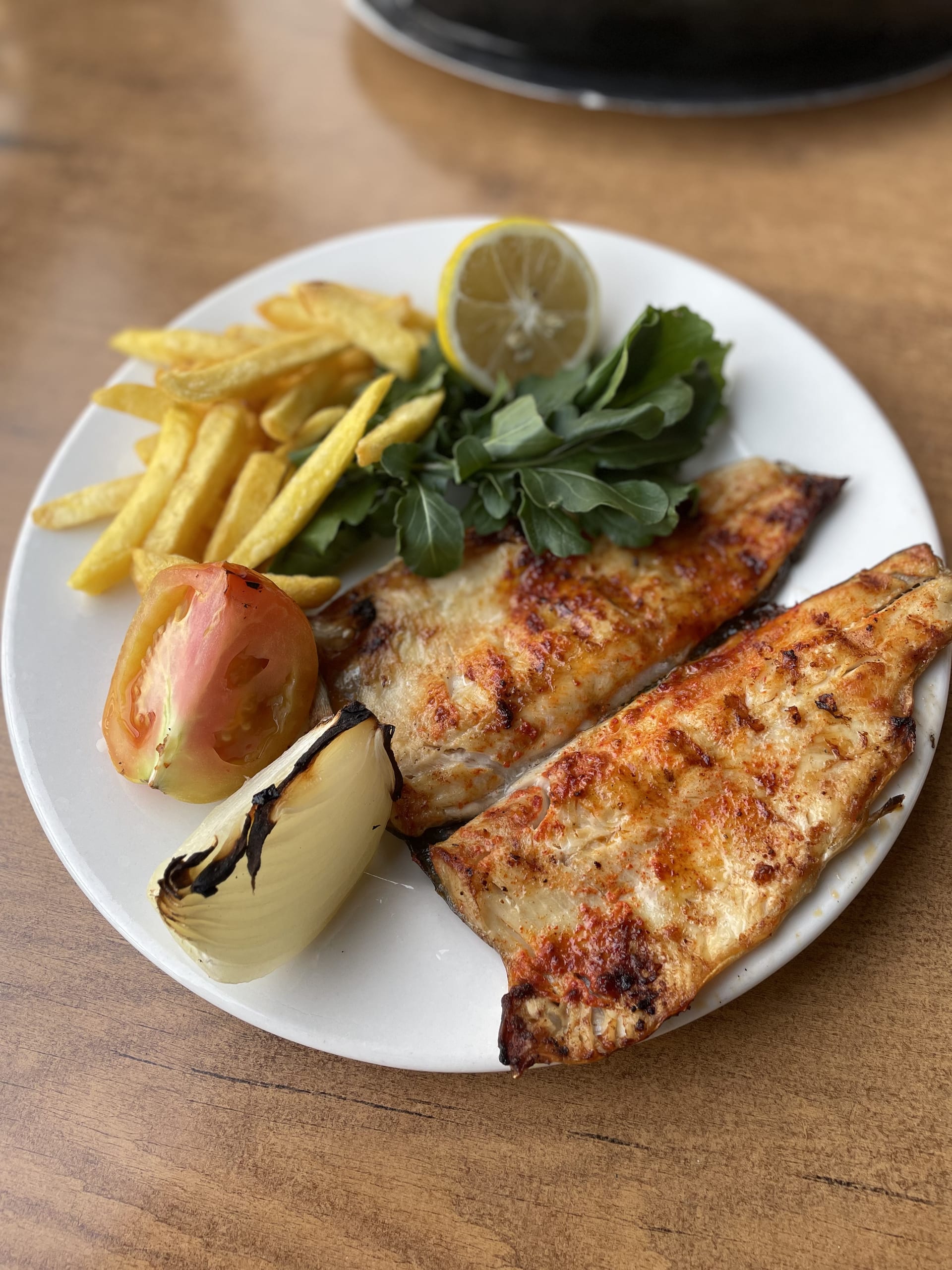 A white plate holds two grilled portions with fries, leafy greens, lemon, tomato and onion.