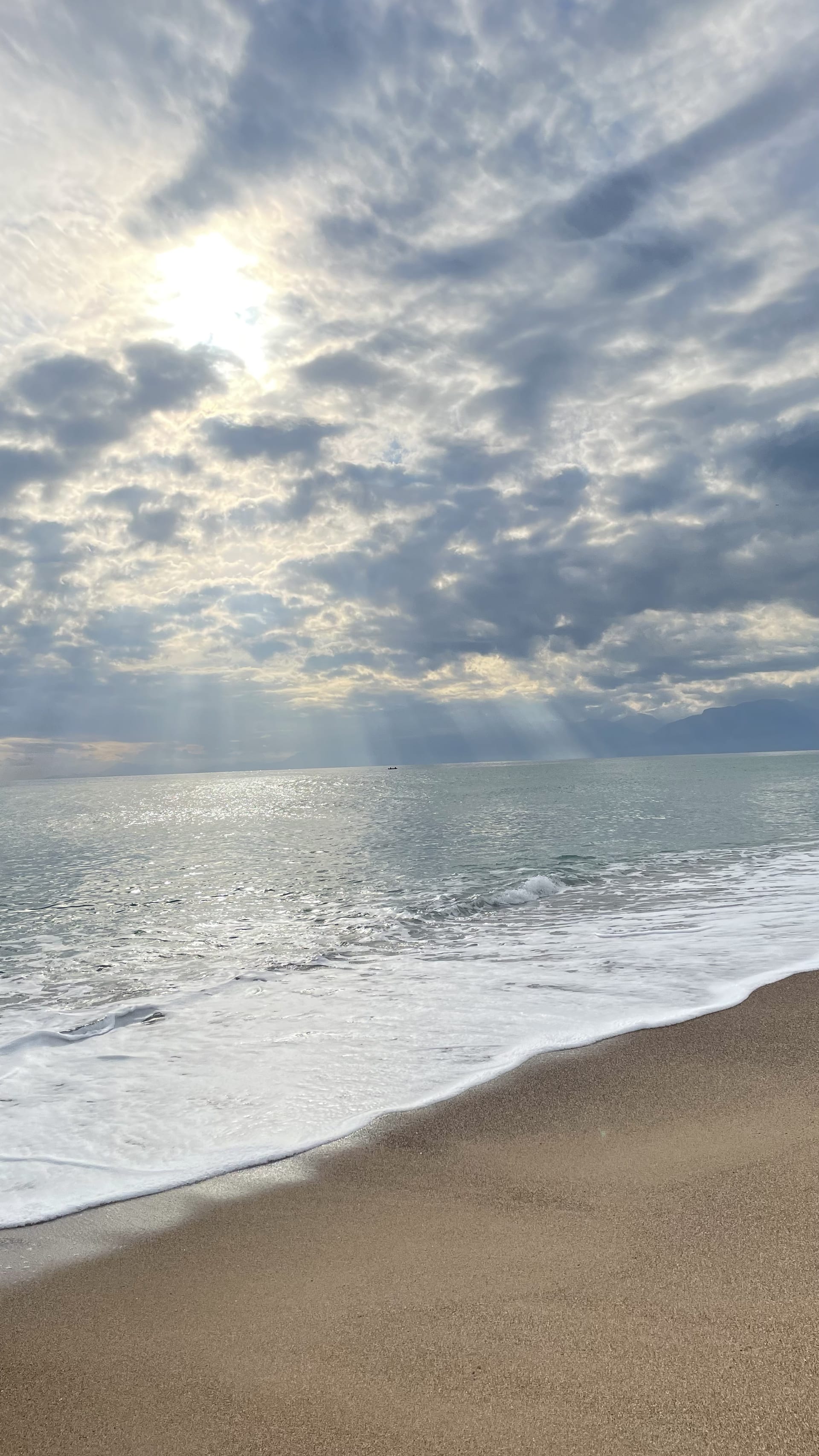 Foamy water reaches a sandy shoreline as sunlight breaks through layered clouds over the sea.