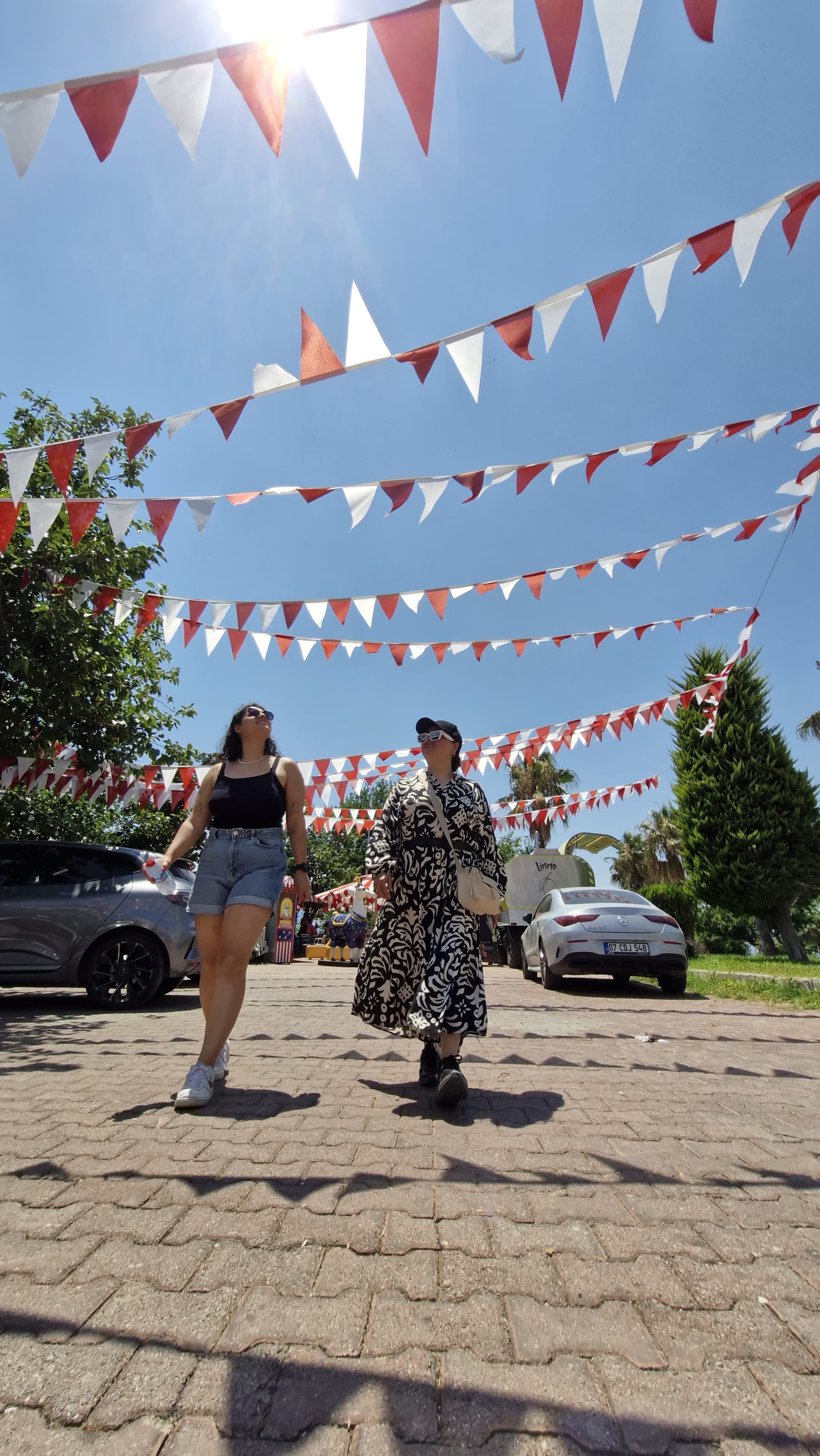 Two women walk beneath rows of red-and-white bunting beside parked cars and greenery.