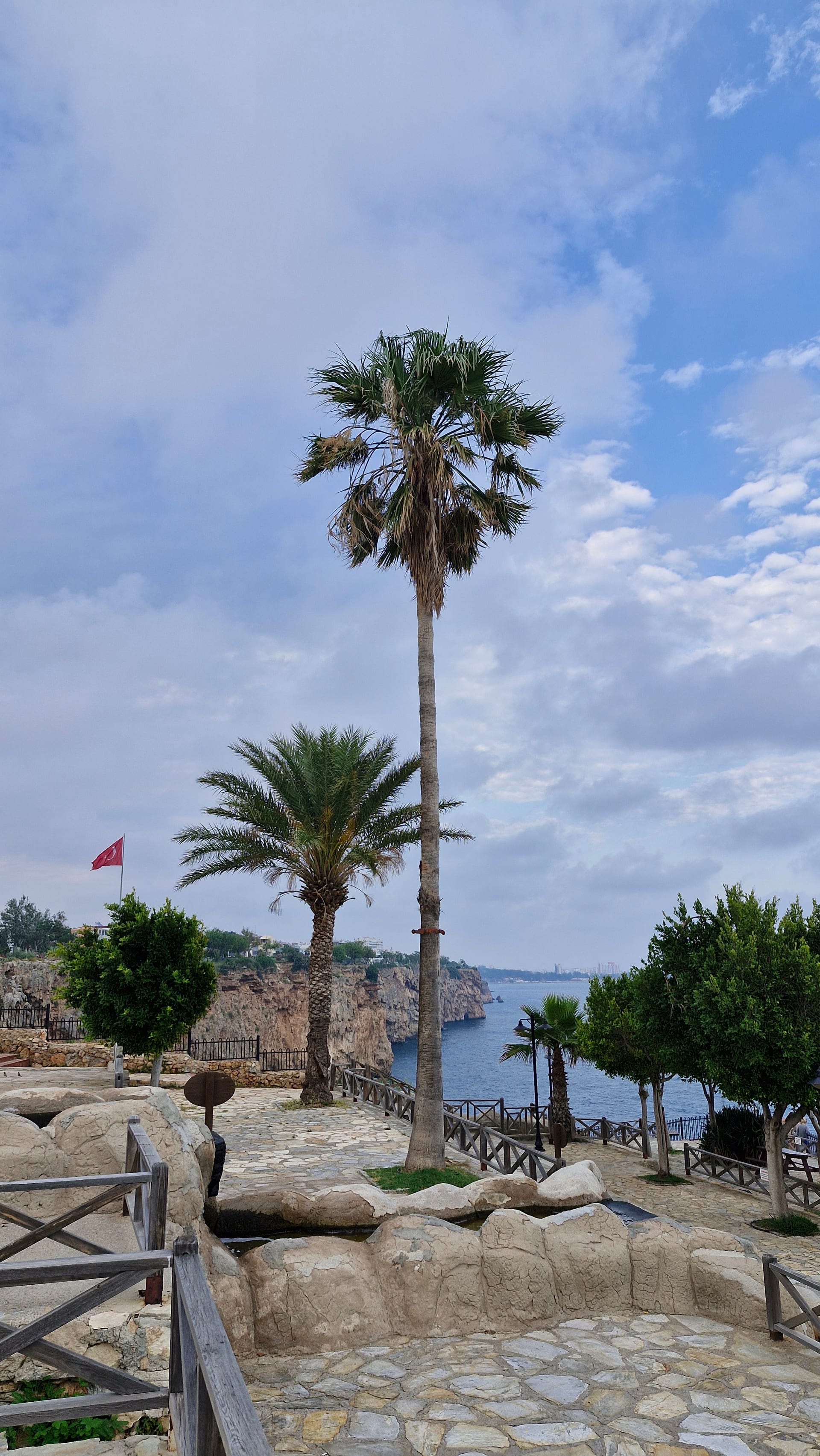 Stone paths, wooden railings and palm trees overlook Antalya’s rocky coastal cliffs and blue water.