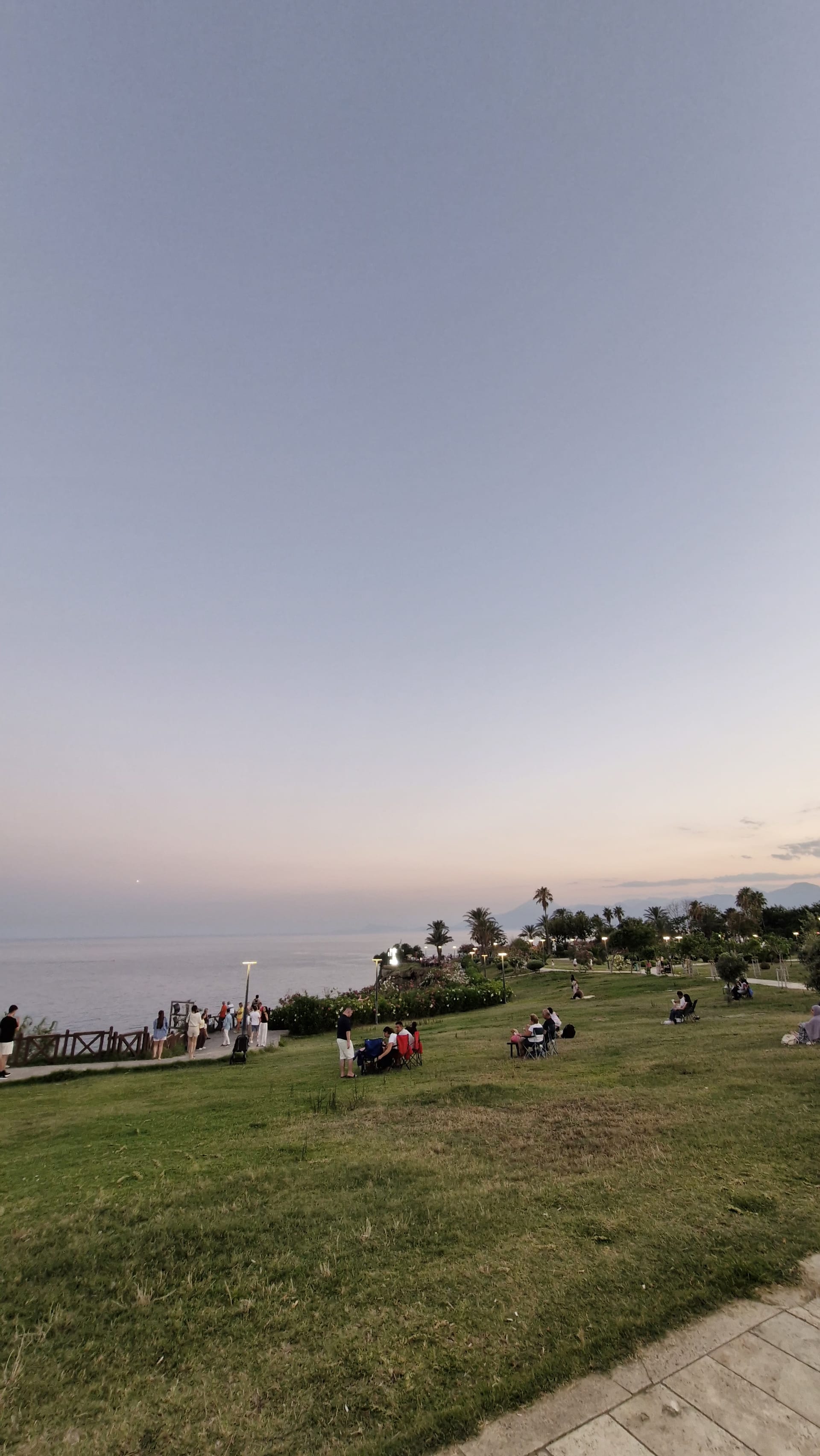 Small groups sit across a broad green lawn beside the sea beneath a pale evening sky.