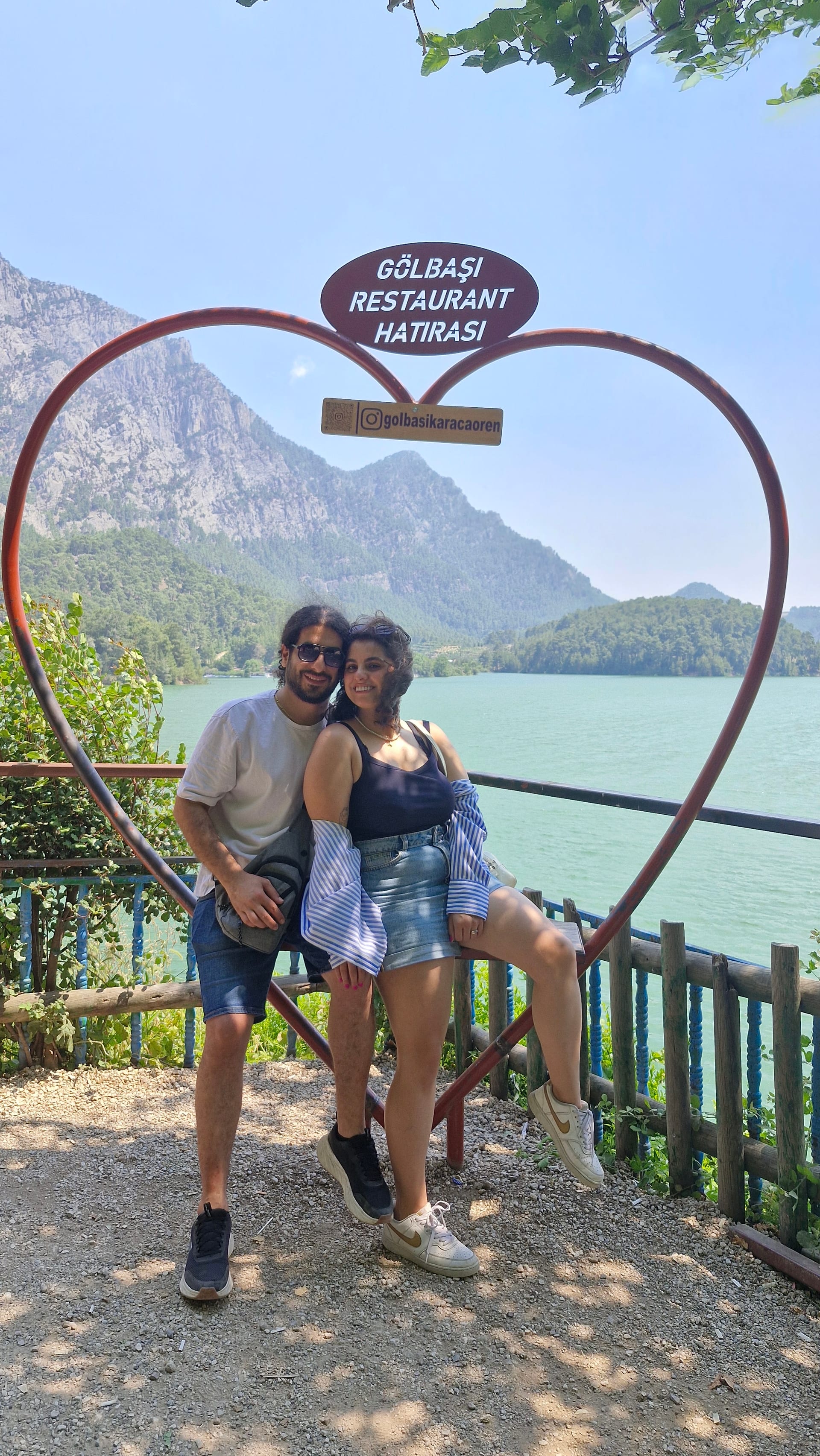 A man and woman pose inside a heart-shaped frame with turquoise water and forested mountains behind them.