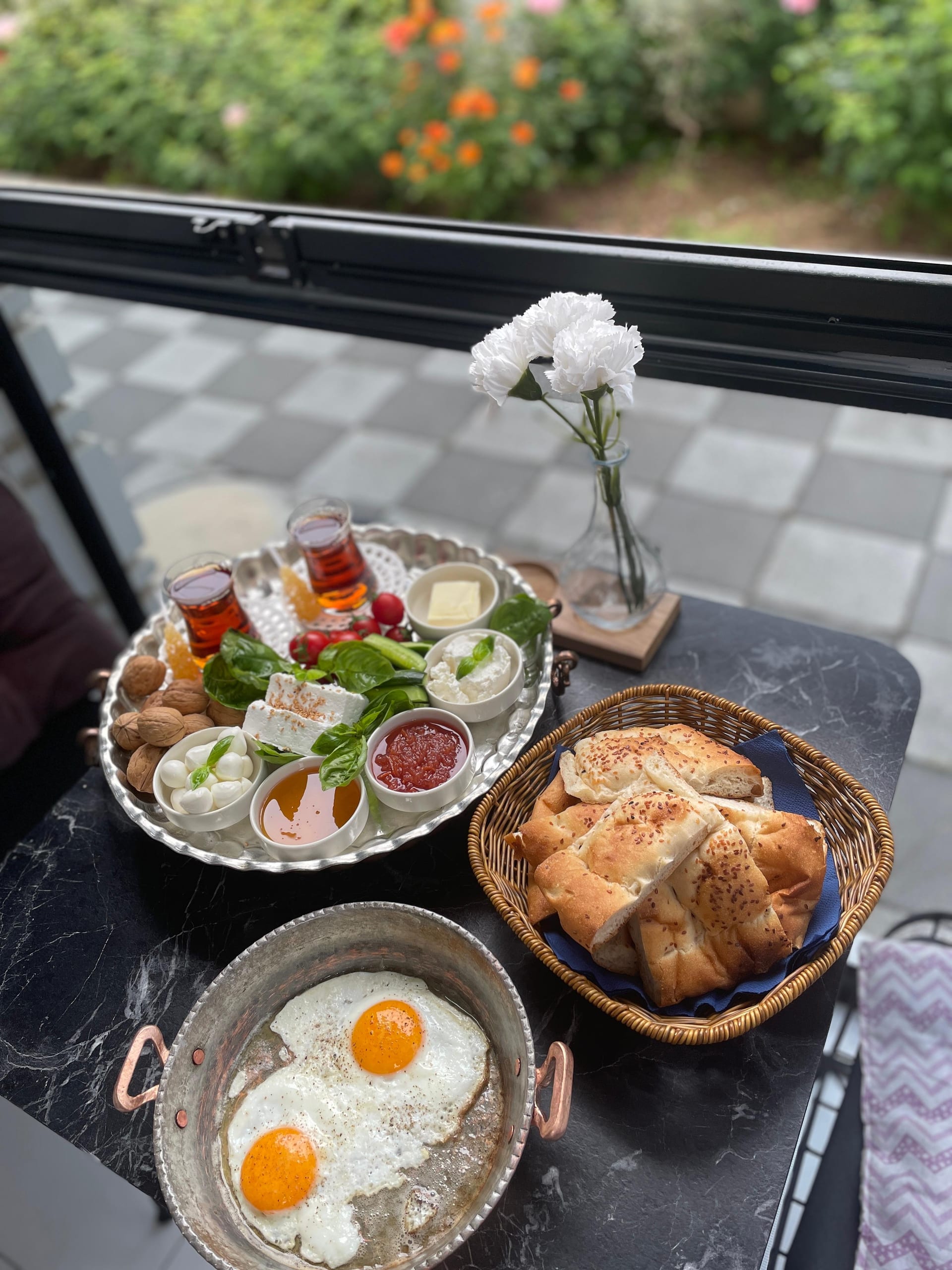 A table beside a window holds glasses of tea, sliced cake, fruit, strawberries, seeds, popcorn, crisps and white flowers.