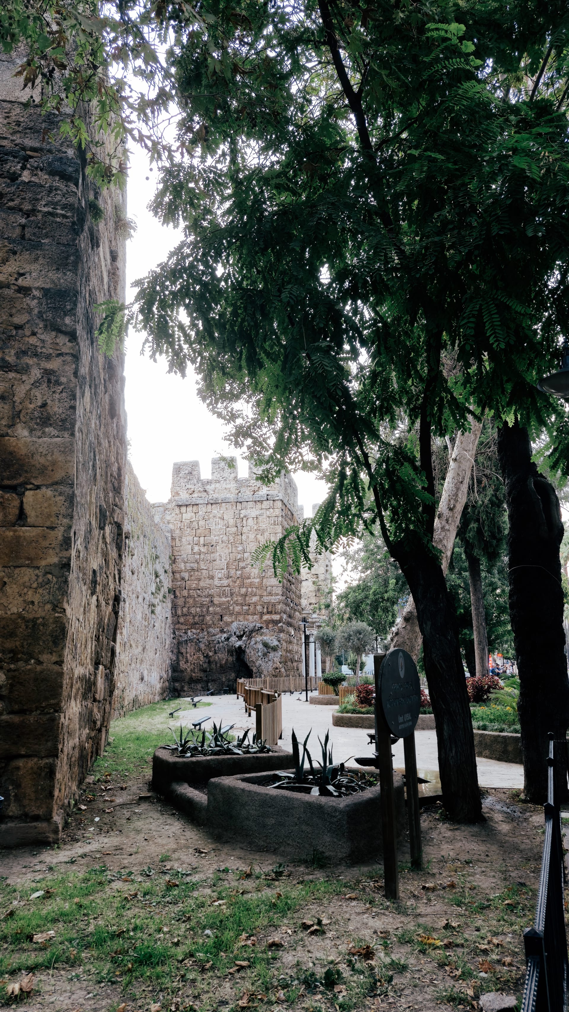 Shaded stone passage with trees, paving and a high old wall.