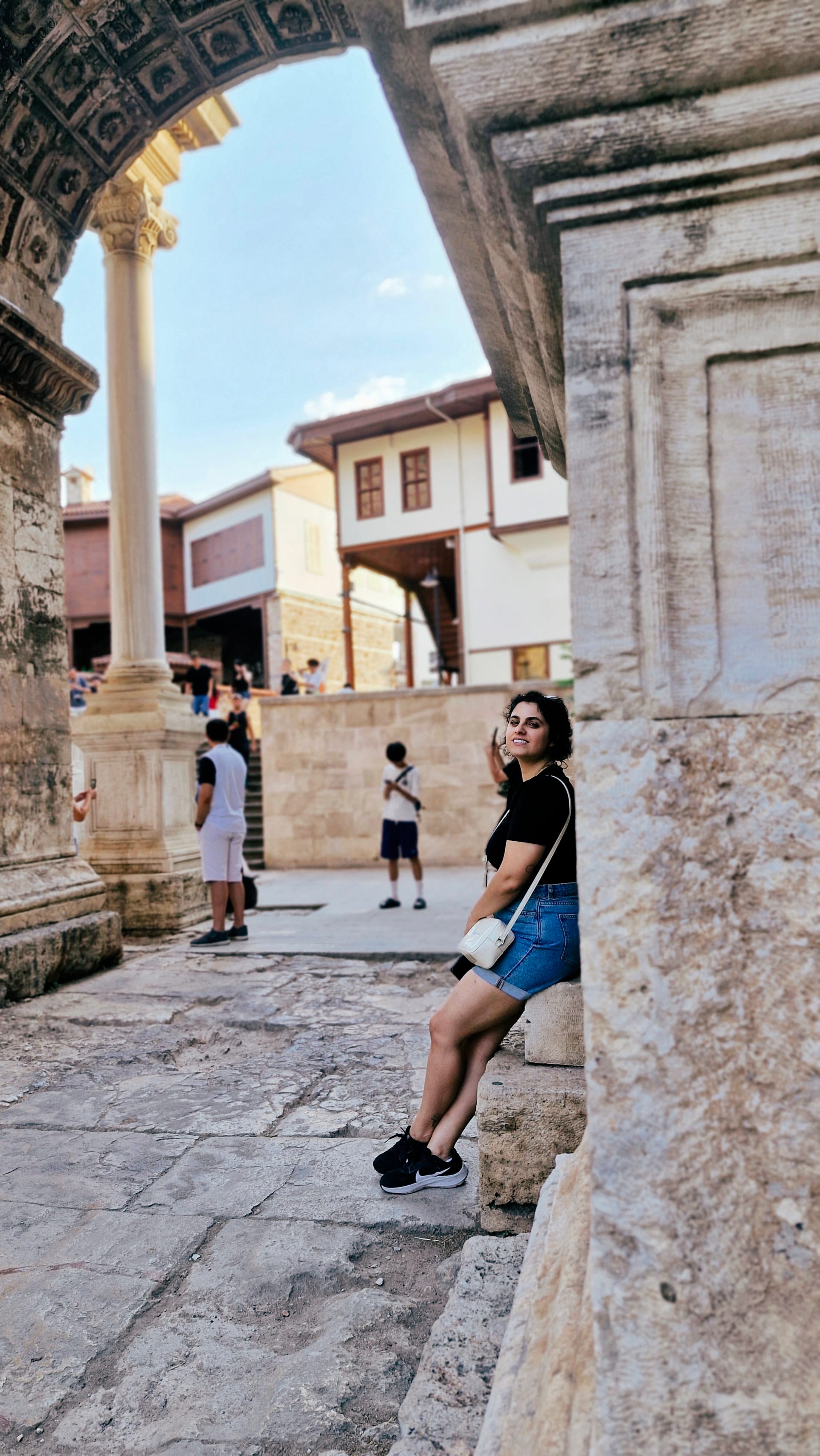 Woman seated on a stone ledge beneath the gate while visitors pass behind her.