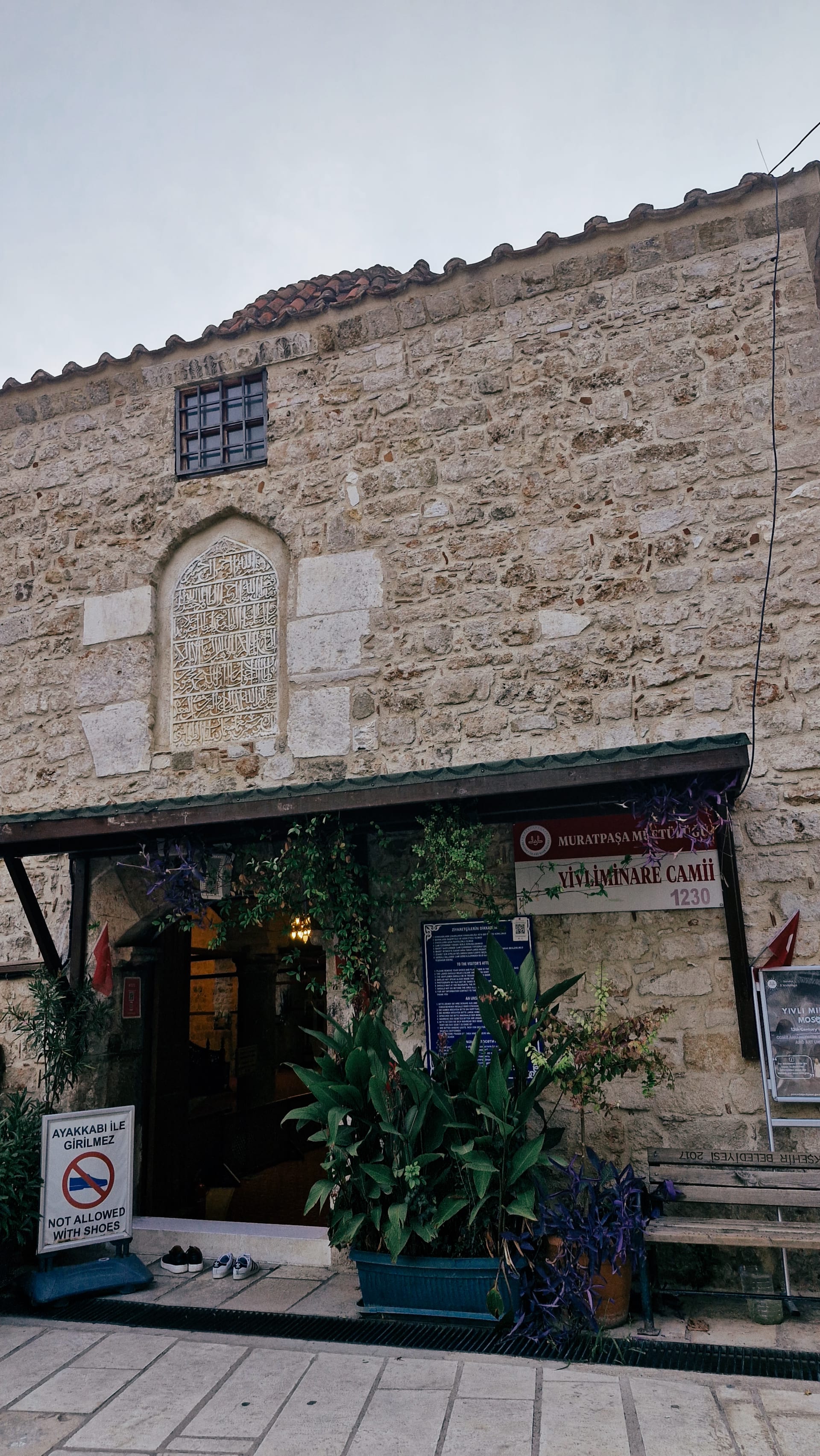 Stone mosque exterior with a small entrance, plants and an inscribed wall panel.
