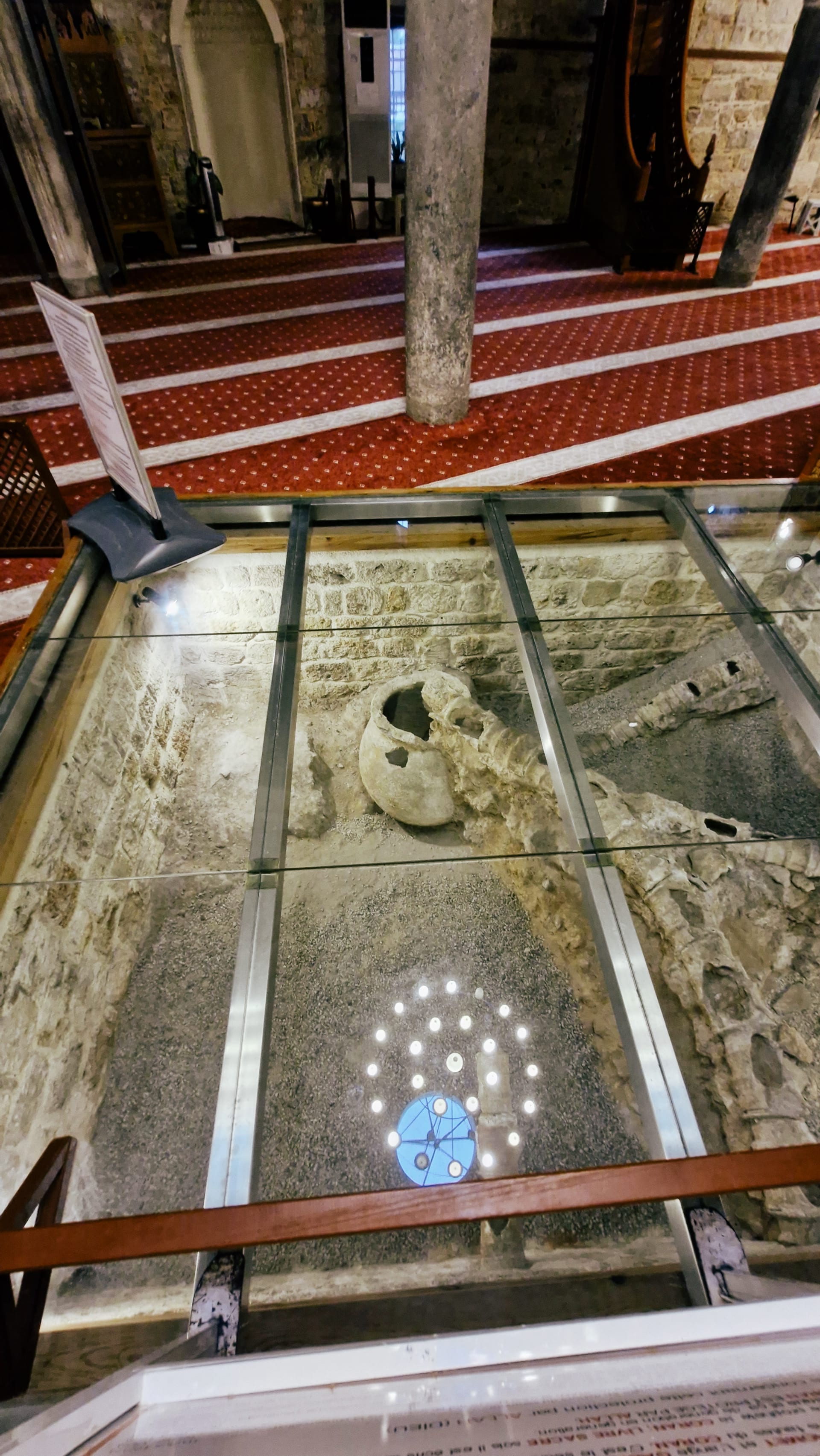 Glass floor revealing stone water channels and a large broken ceramic vessel below.