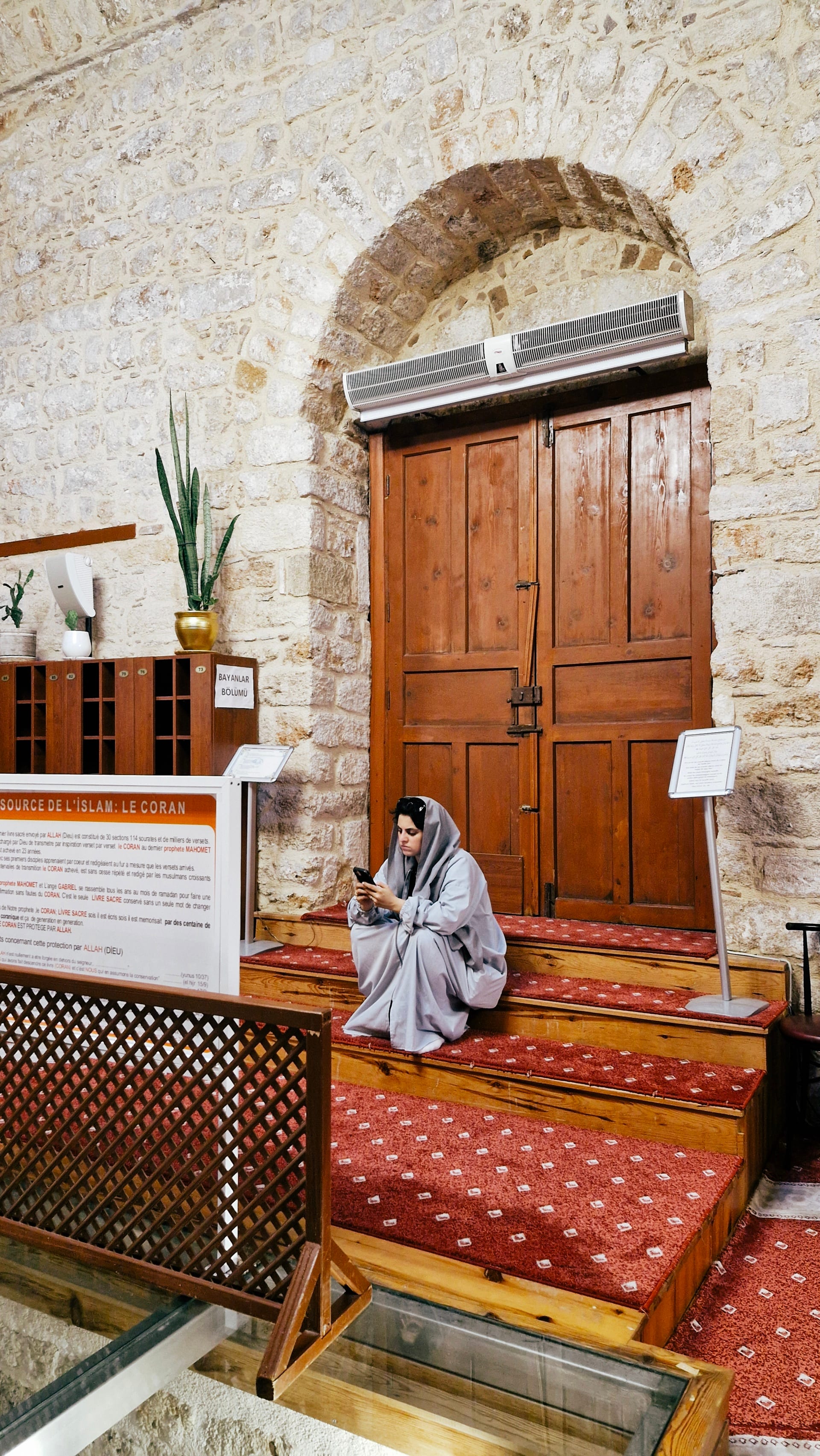 Woman sitting beside a tall wooden door on the red mosque carpet.