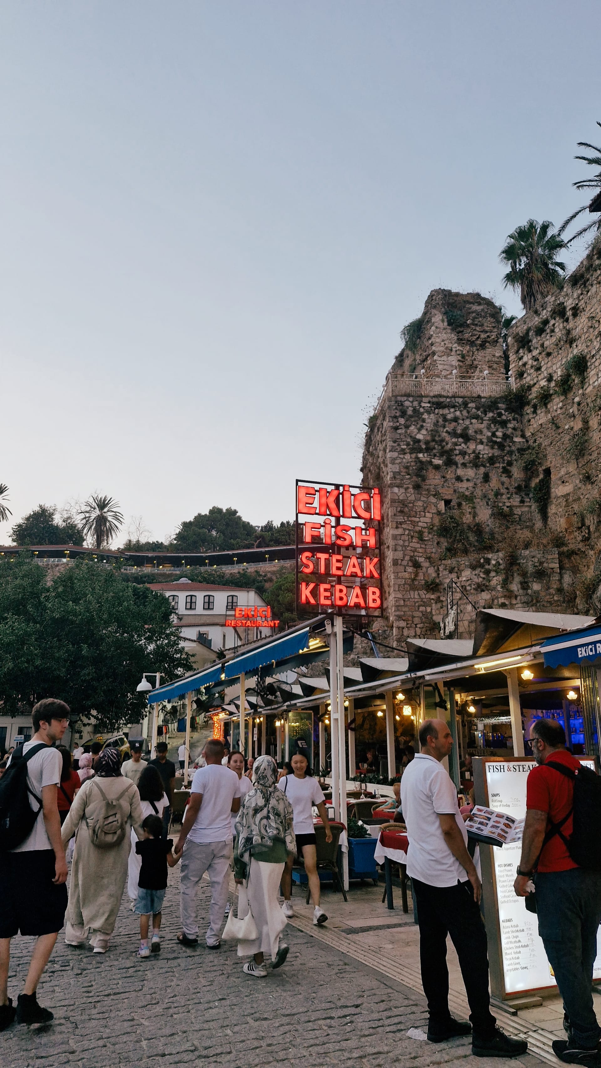 Crowded harbor walkway beside restaurants, boats and a rough stone wall.