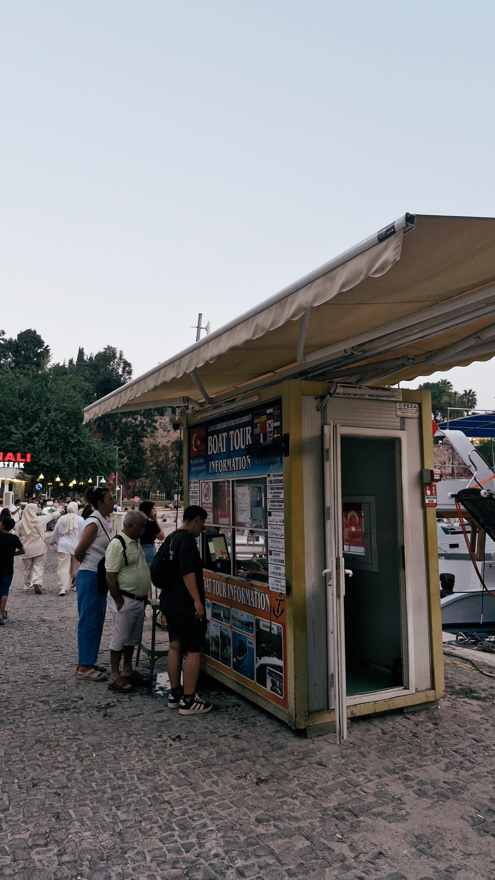 Visitors gathered around a small harbor ticket booth beside moored boats.