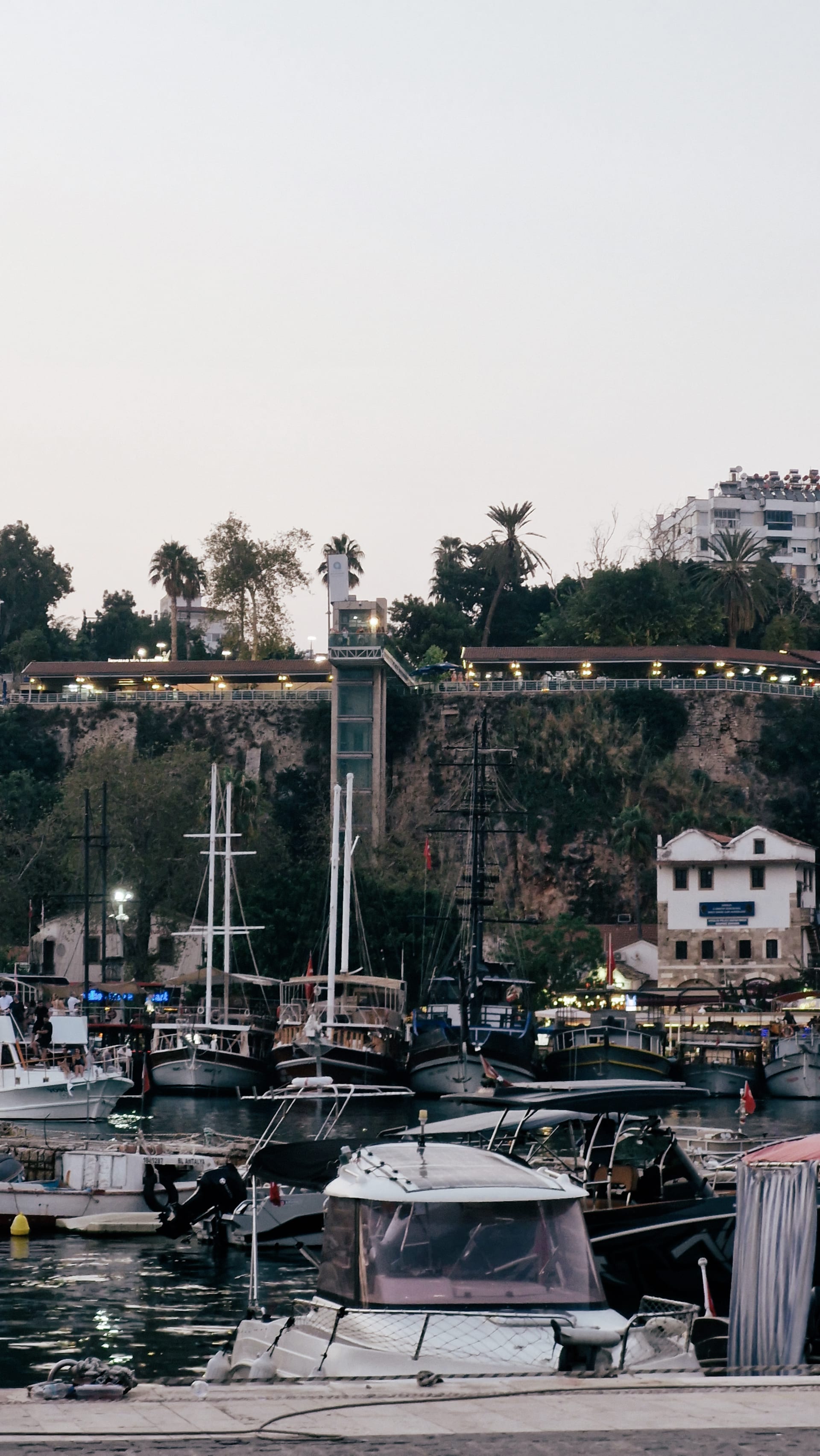 Boats packed below high stone cliffs topped with trees and buildings.