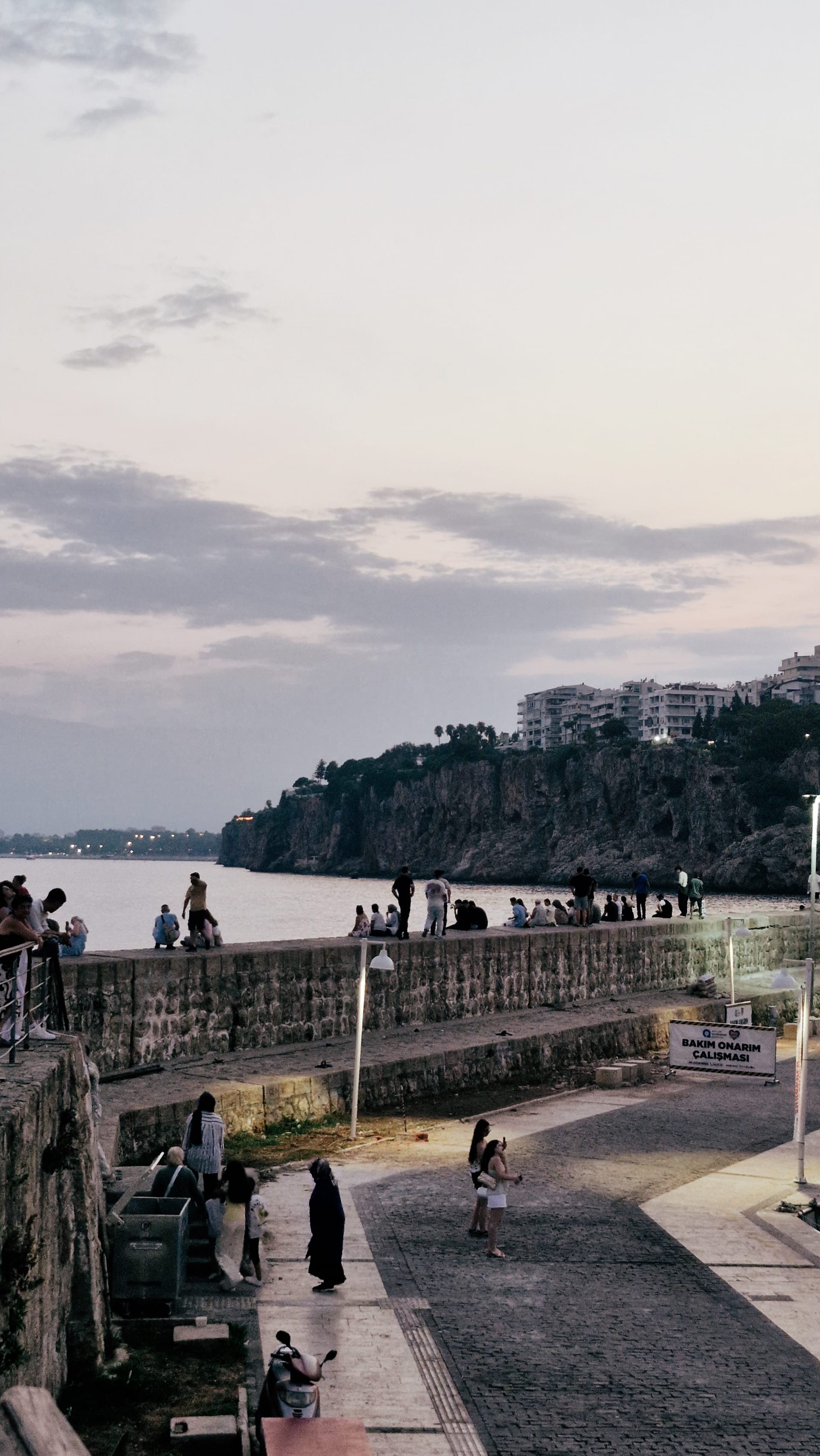 Stone promenade curving around the water below Antalya's cliffs at dusk.