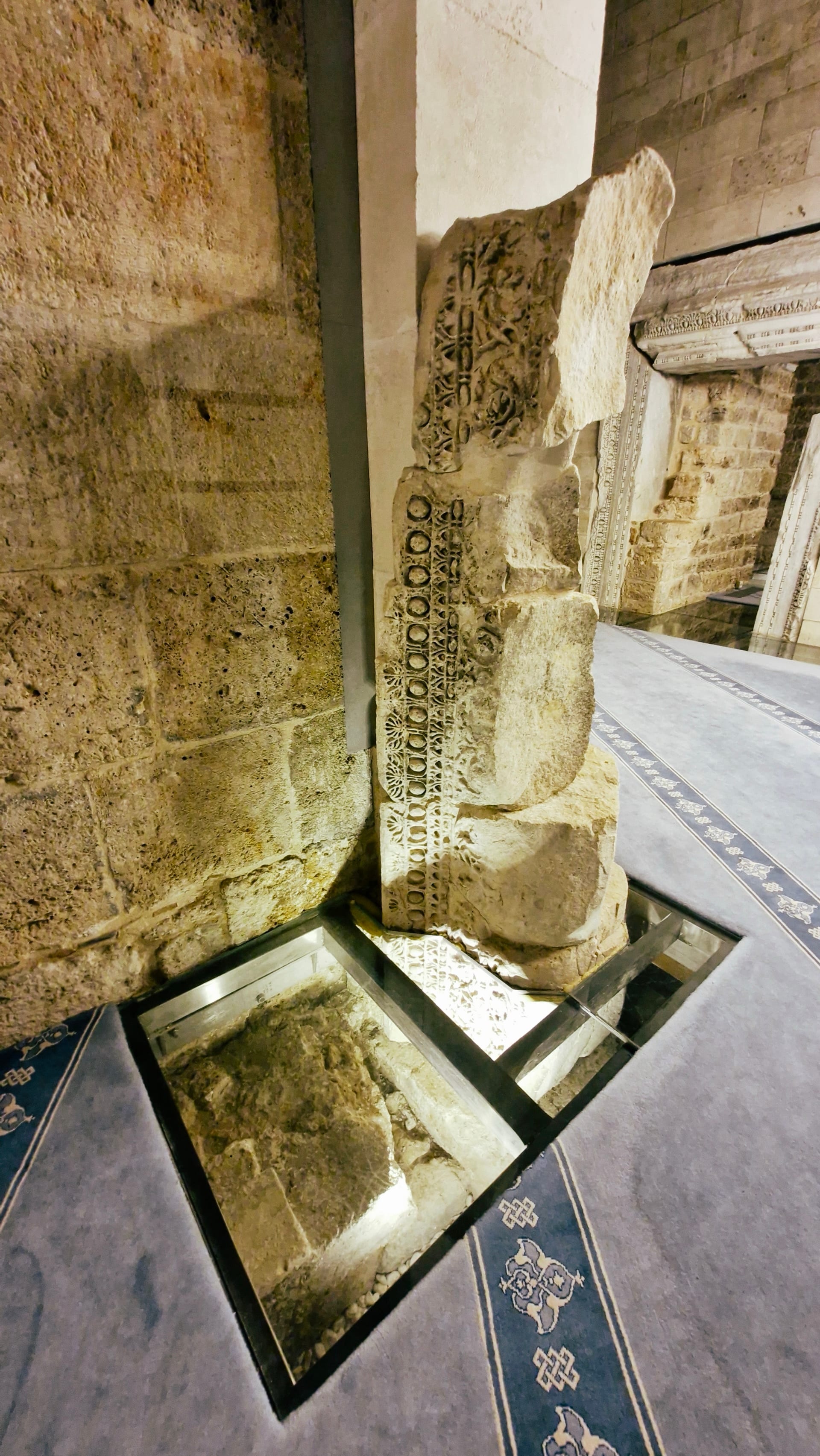 Carved upright stone fragment beside a lit glass floor panel.