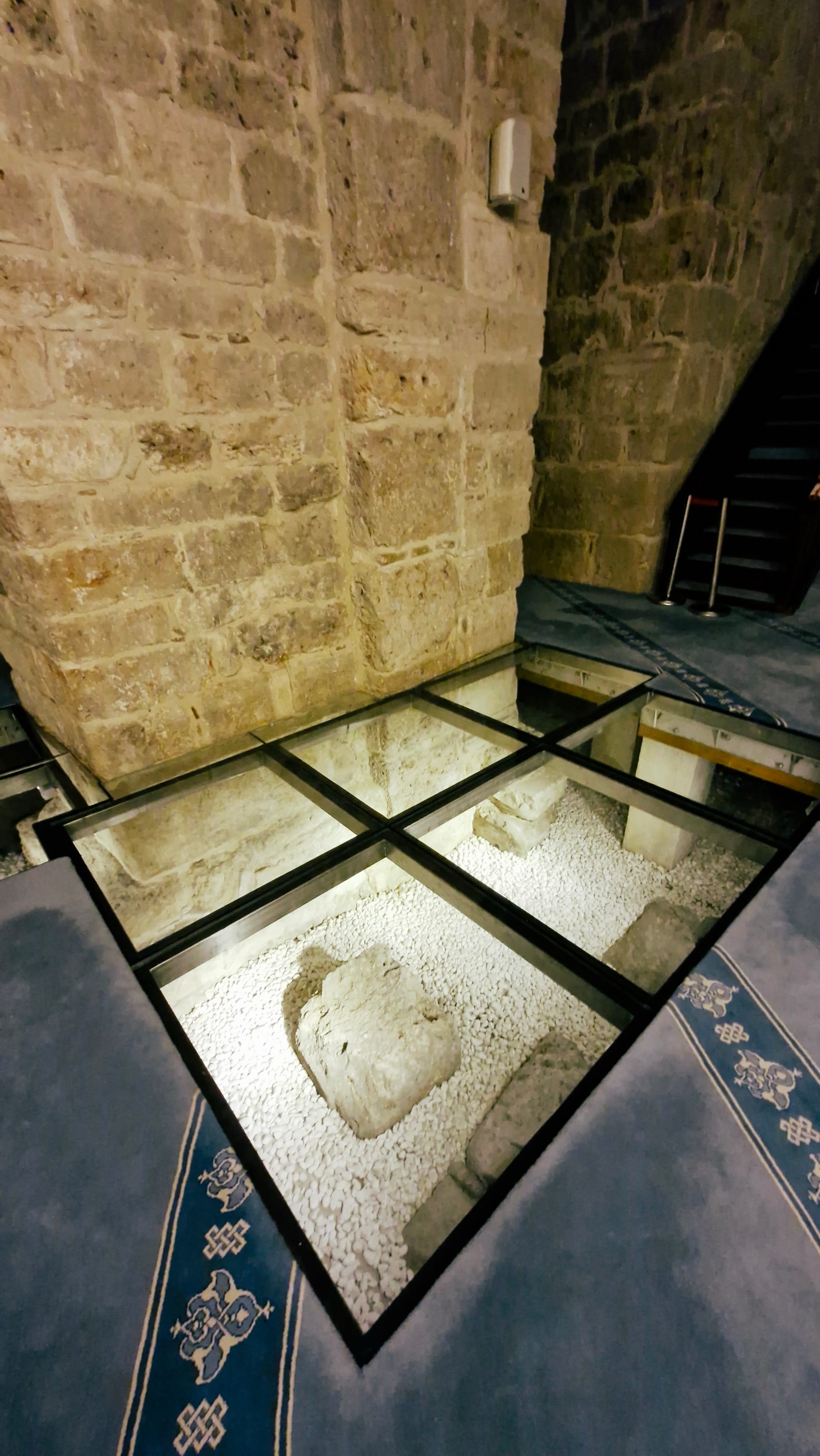 Corner of glass flooring revealing stone blocks and a round fragment below.