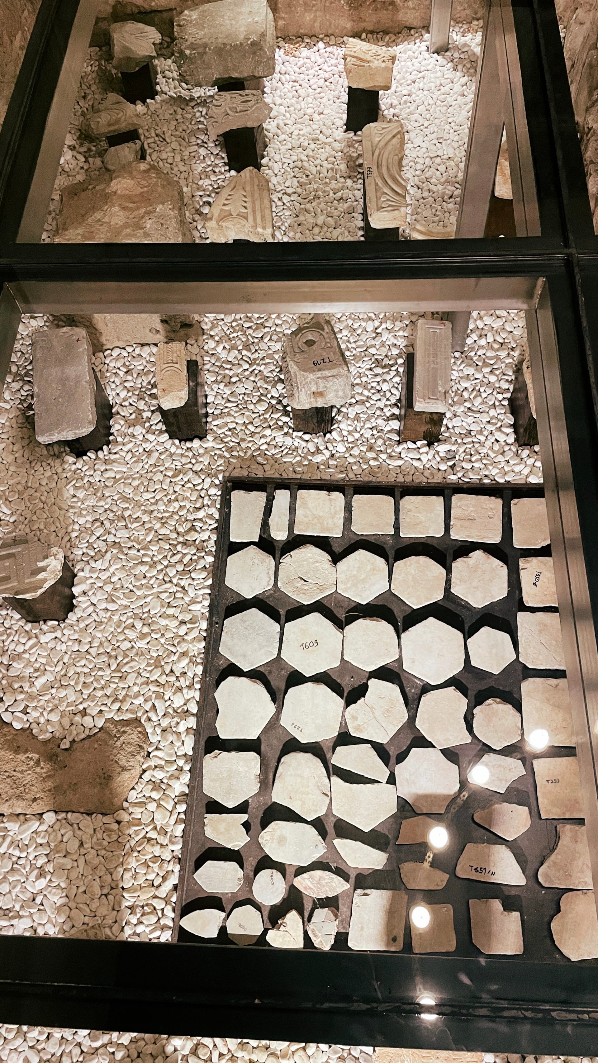 Carved and inscribed stone fragments arranged beneath glass on white gravel.