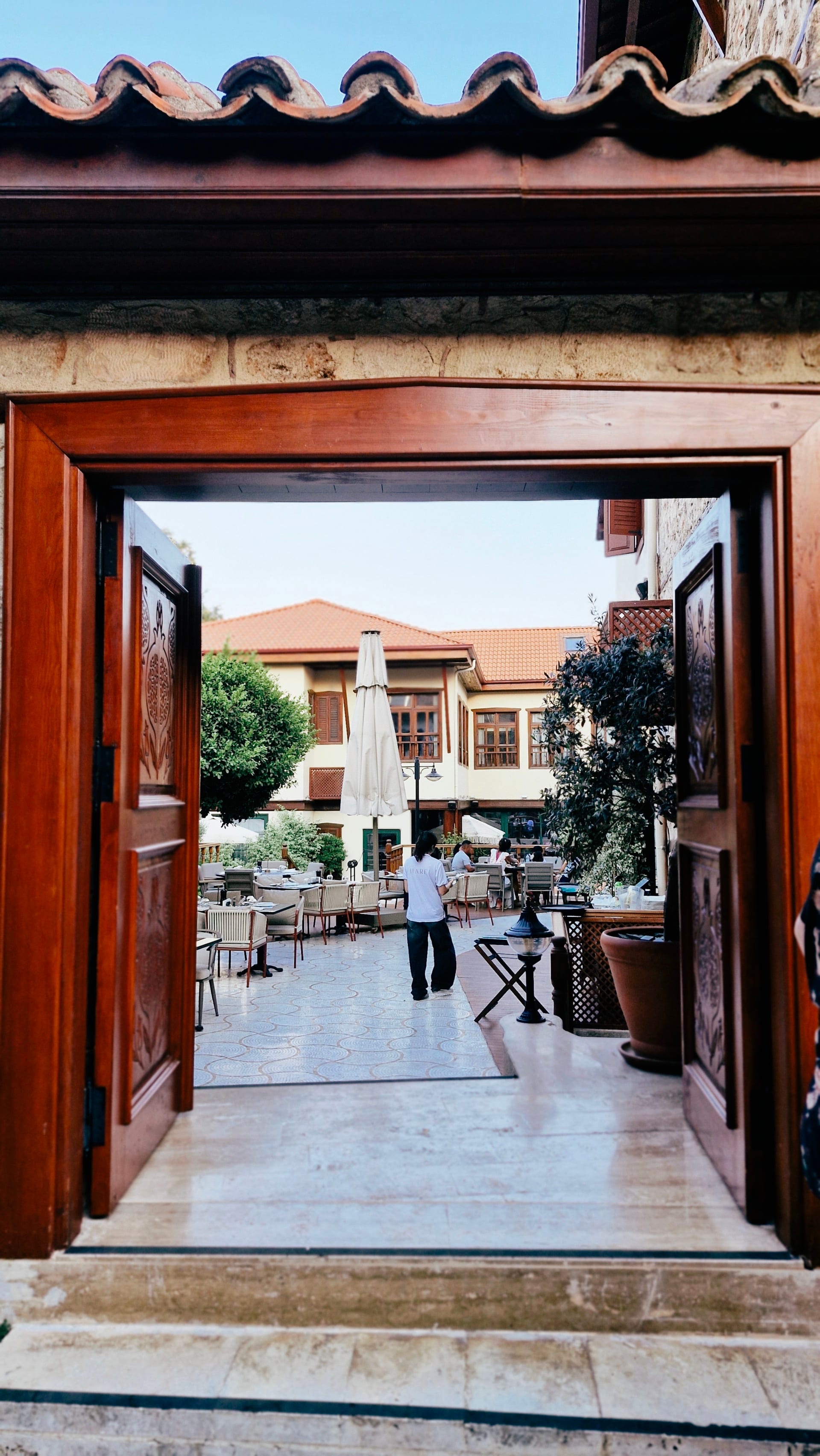 An open wooden doorway frames a courtyard with café tables and a white parasol.