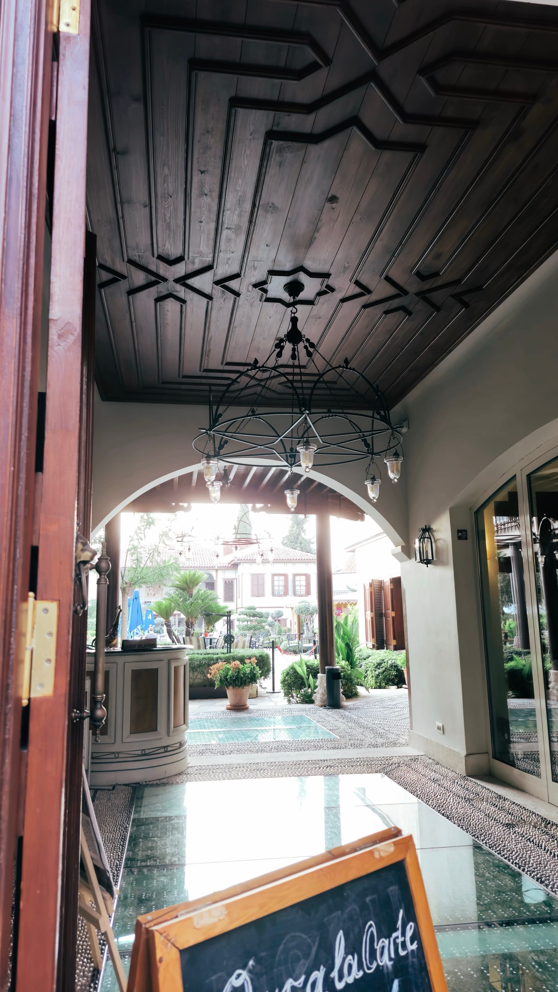 A chandelier hangs from a patterned passage leading to a green courtyard.
