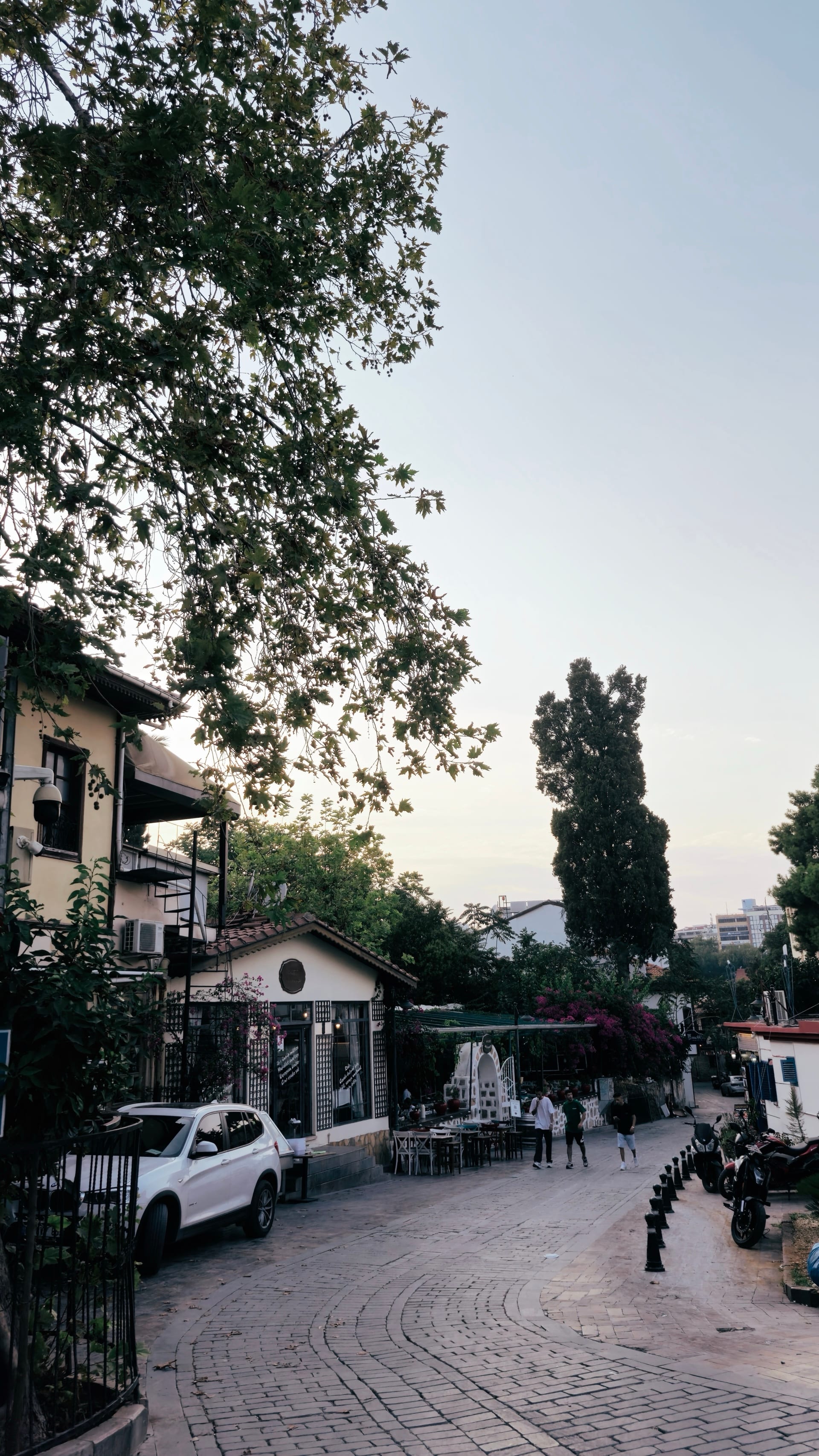 A curving cobbled lane descends past trees, café tables, parked vehicles, and pedestrians.