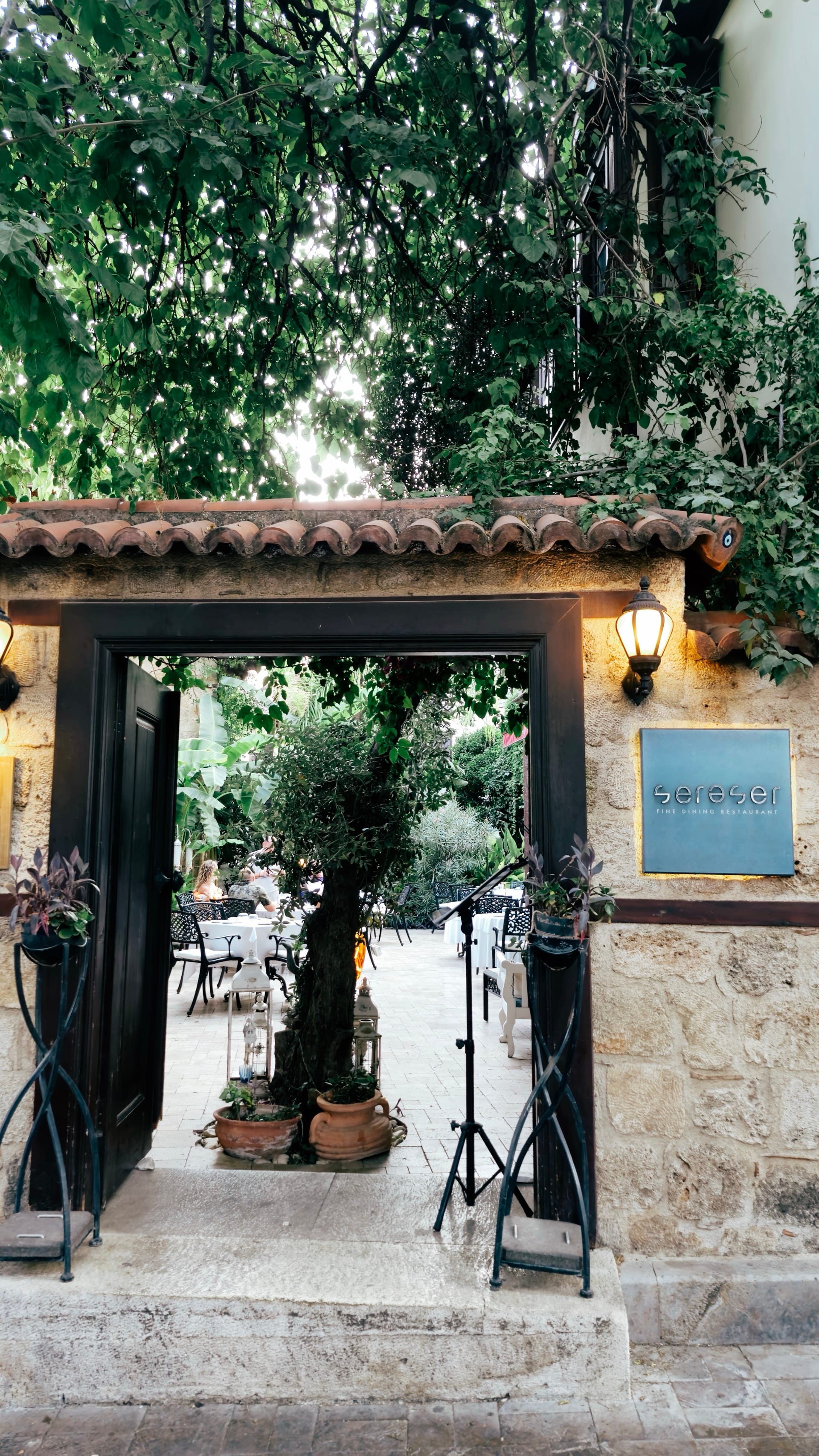 A tiled stone gateway opens into a leafy garden with tables.