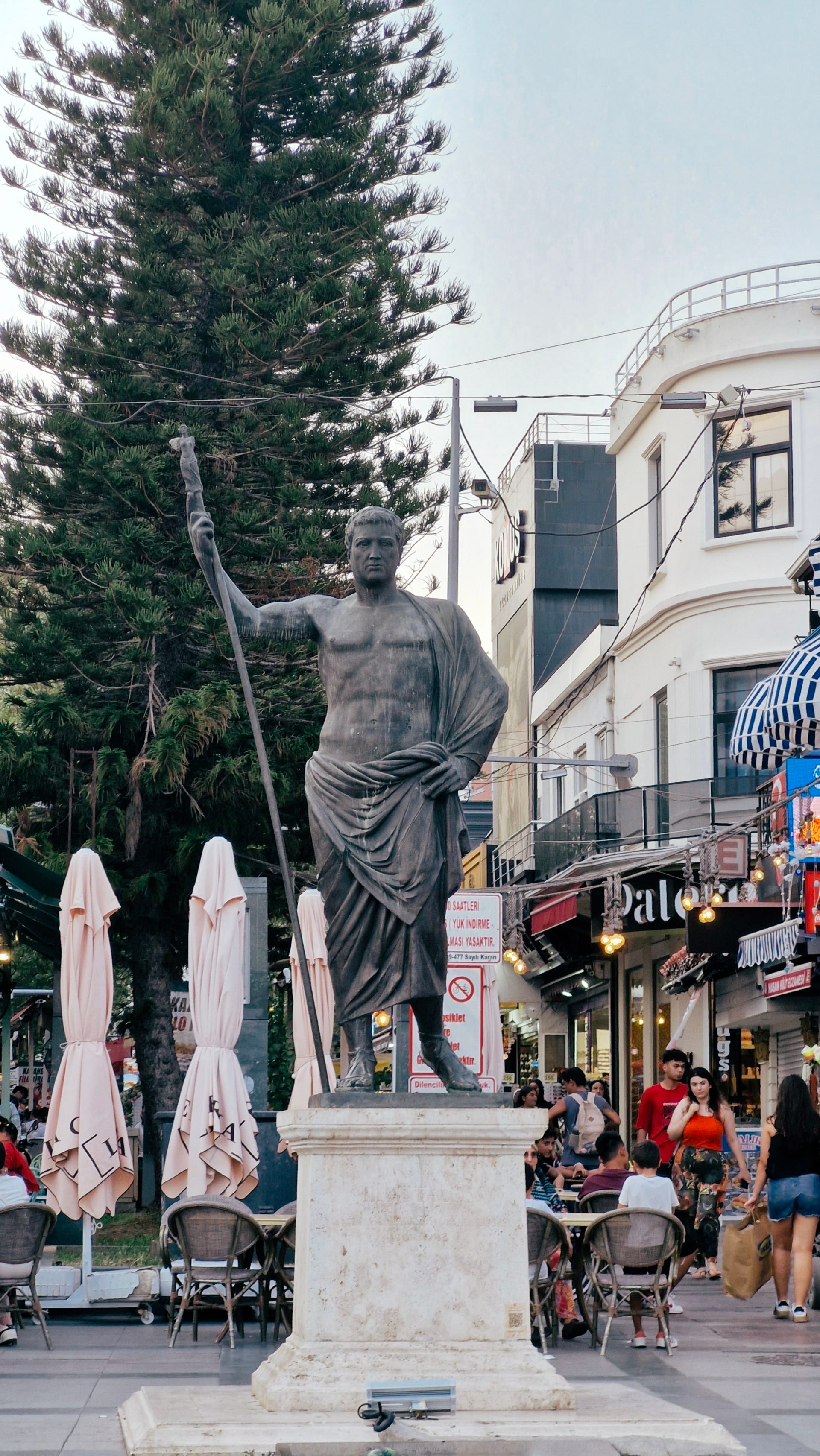 A robed statue holding a long staff stands among café tables and shops.