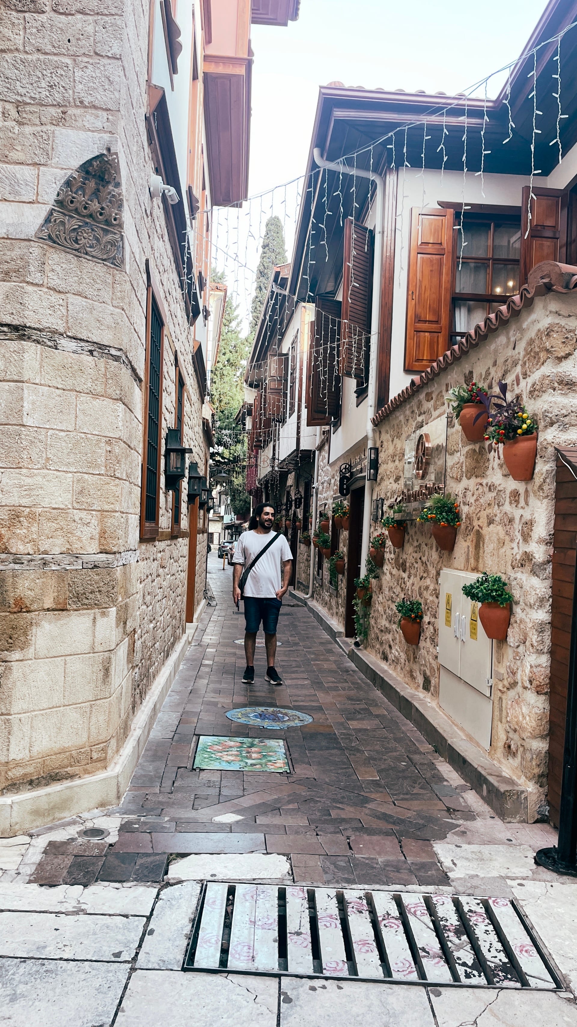 A man in a white shirt walks along a narrow lane decorated with flowers and mosaics.