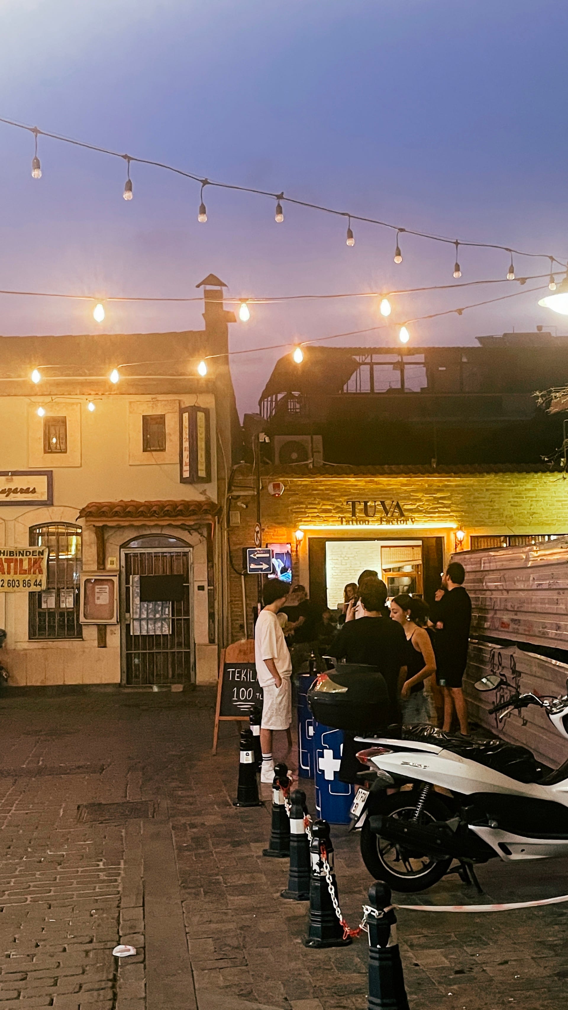 Diners gather under string lights on a night street with scooters parked nearby.