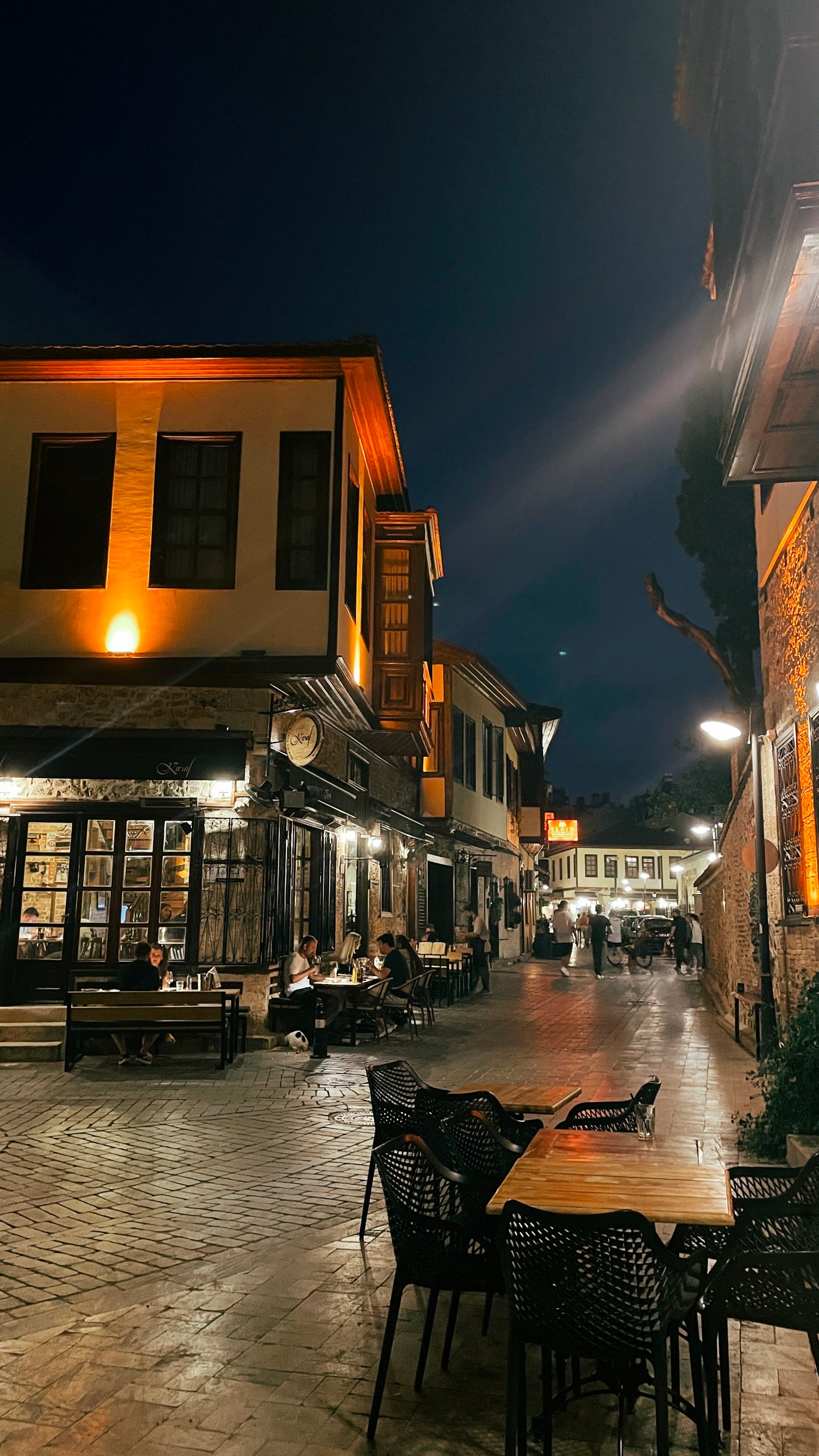 Empty café tables line a wet stone street reflecting lights at night.