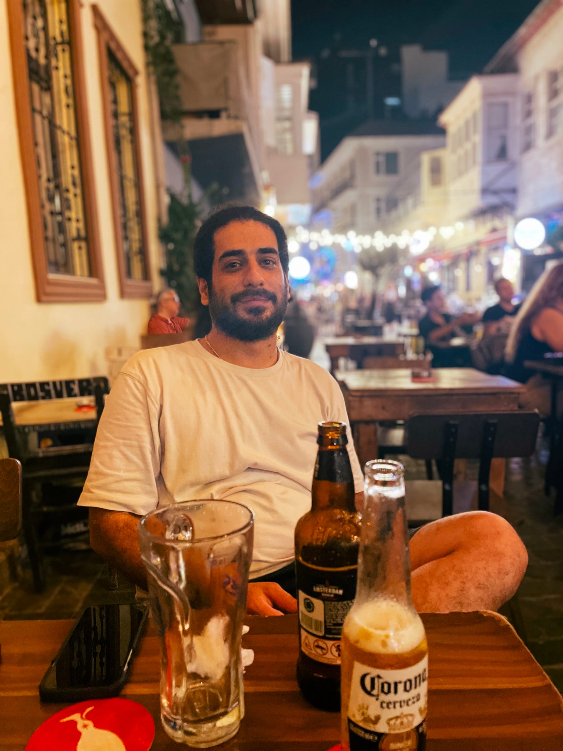 Man seated at an outdoor table with a beer bottle and empty glasses.
