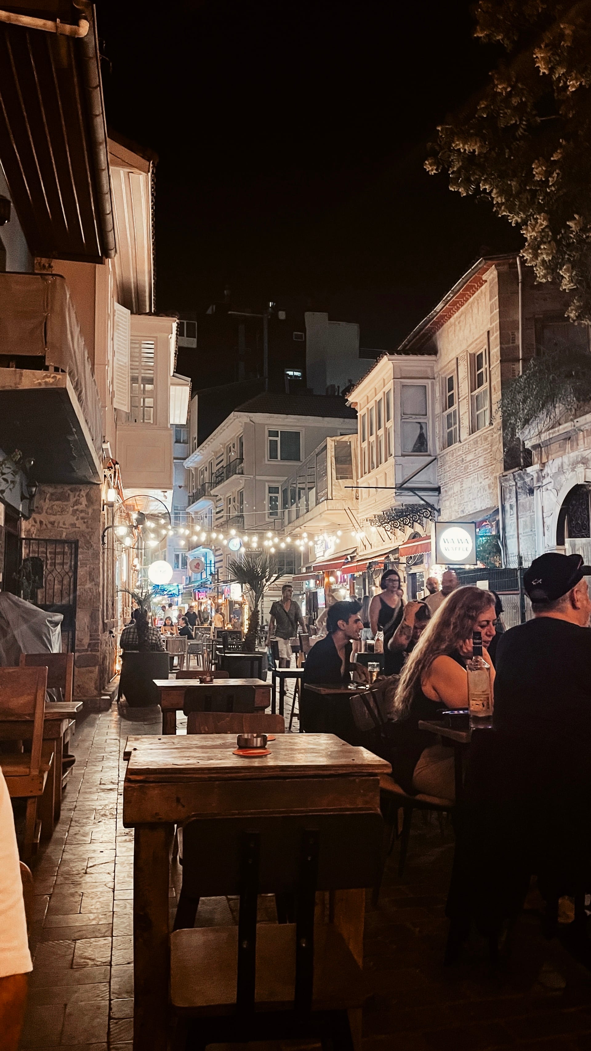 Outdoor café tables facing a brightly lit, crowded old-town street at night.