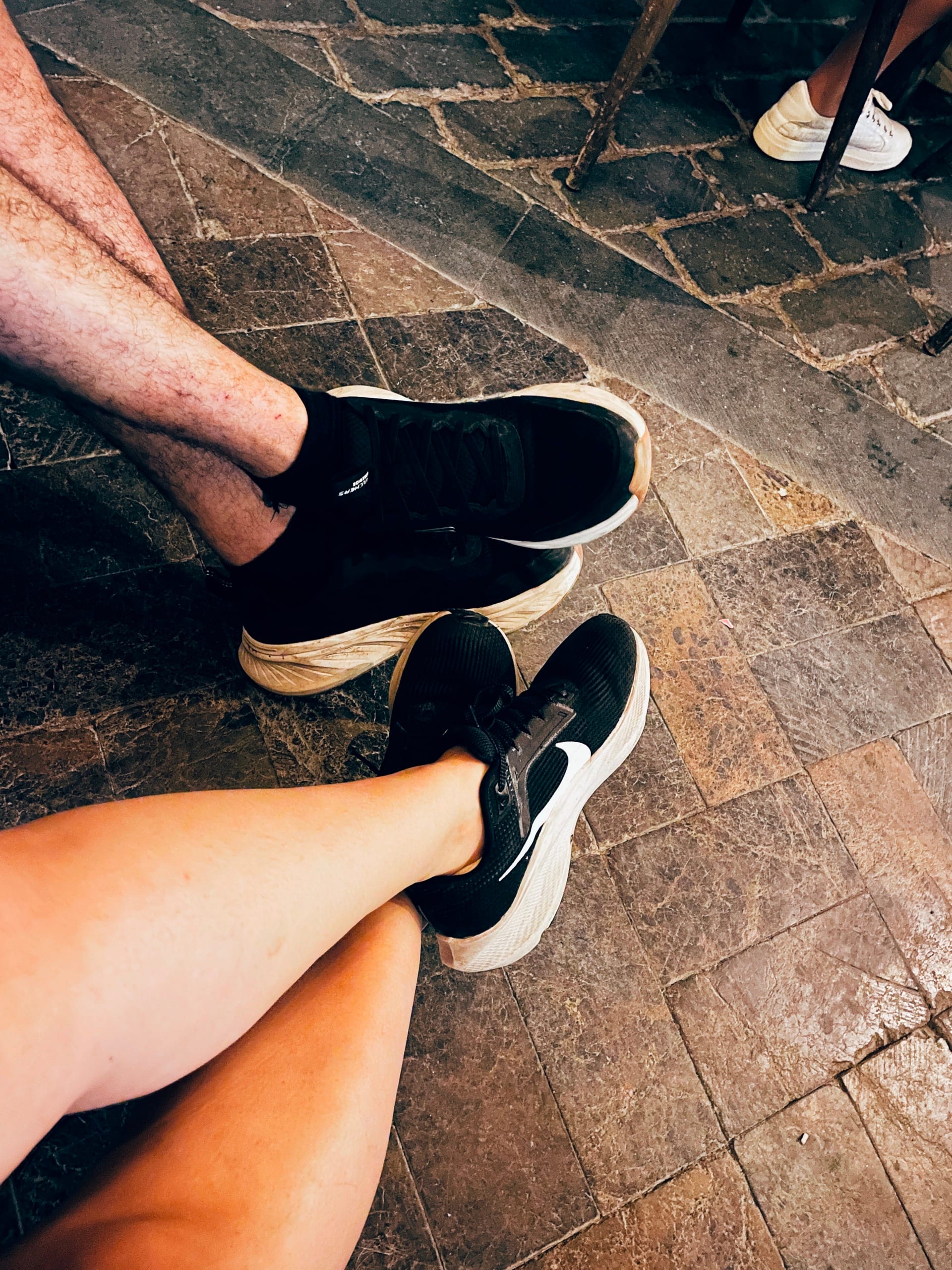 Two pairs of black shoes and crossed legs are seen above a stone pavement.